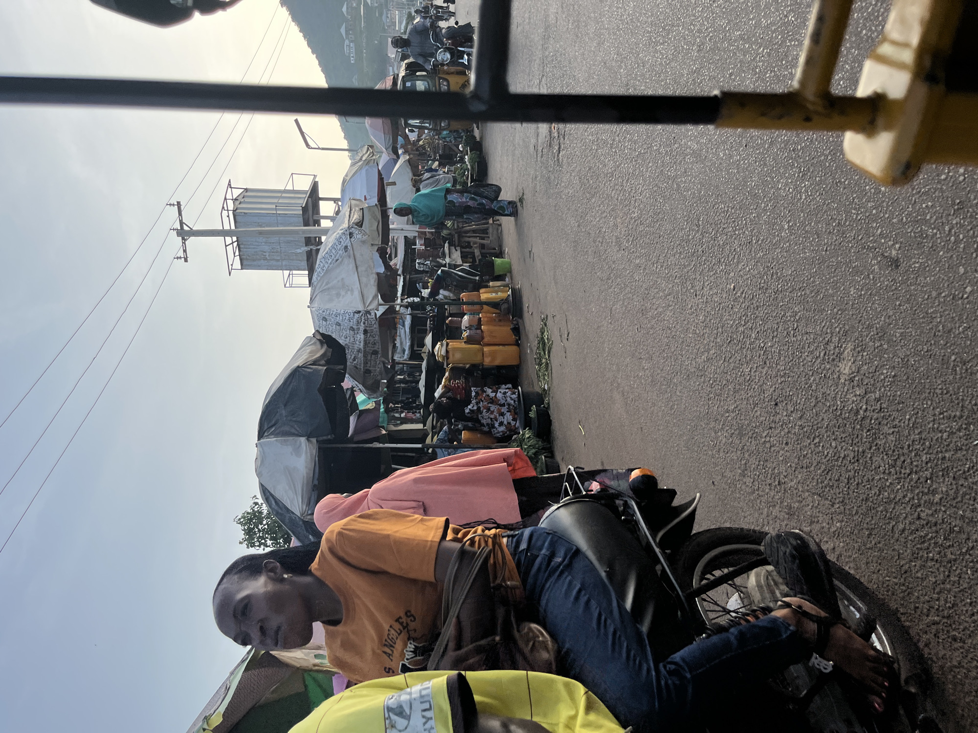 A low-angle, full shot captures a street market scene in Unknown, Unknown, at dusk. In the foreground, a woman with dark skin and braids, wearing an orange t-shirt that reads "Los Angeles" and blue jeans, sits on a motorcycle. She is looking to her left with her mouth slightly open, as if speaking. To her left, a person in a yellow shirt is partially visible. Behind the woman, a person in a pink hijab is seen. The background reveals a bustling market with numerous stalls, some covered by umbrellas. Stacks of yellow jerrycans are prominent. Several people are walking and riding motorcycles. A utility pole with wires and a water tower are visible in the distance, with a hillside and houses in the far background. The sky is clear and pale.