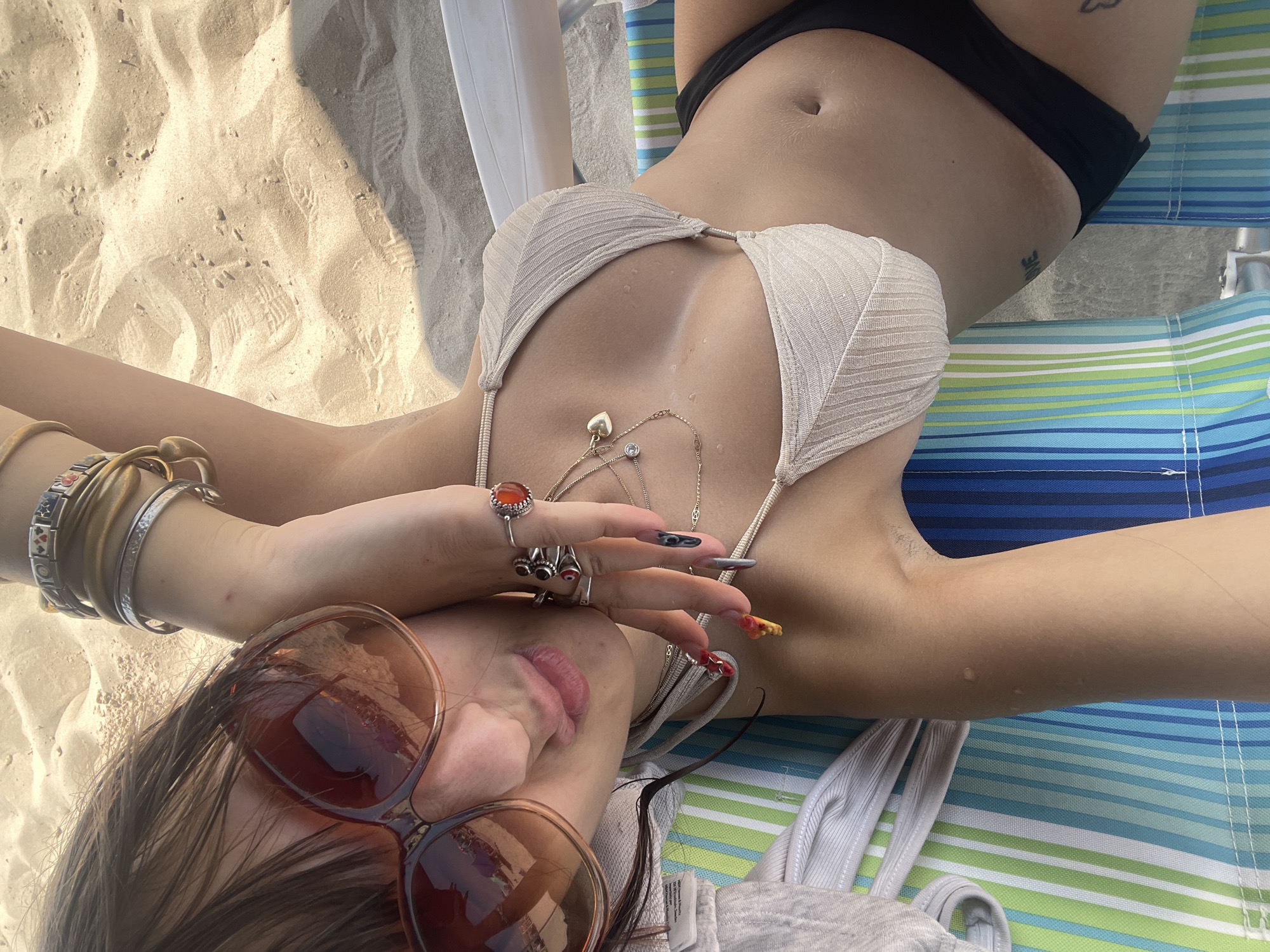 A woman is lying on a beach chair on a sandy beach. She is wearing a light-colored bikini top and black bikini bottoms. She has on large sunglasses, several rings on her fingers, and bracelets on her wrist. The beach chair has blue, green, and white stripes. The sand is light in color and has footprints on it. The woman's skin is glistening, suggesting she may be wet. The lighting indicates it is daytime. The location is the Hof HaCarmel Regional Council, Israel.