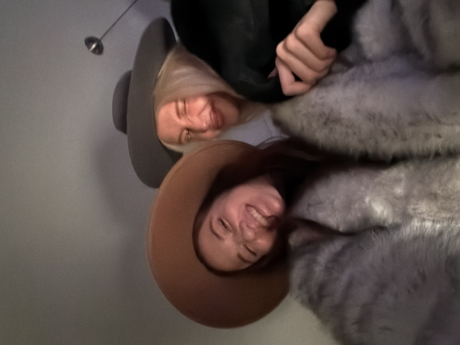 Two women wearing hats and furry coats are angled close to the camera. The woman on top has blond hair and a gray hat. The woman on the bottom has brown hair and a tan hat. Both are smiling. A dark jacket is visible. A light fixture hangs in the background.