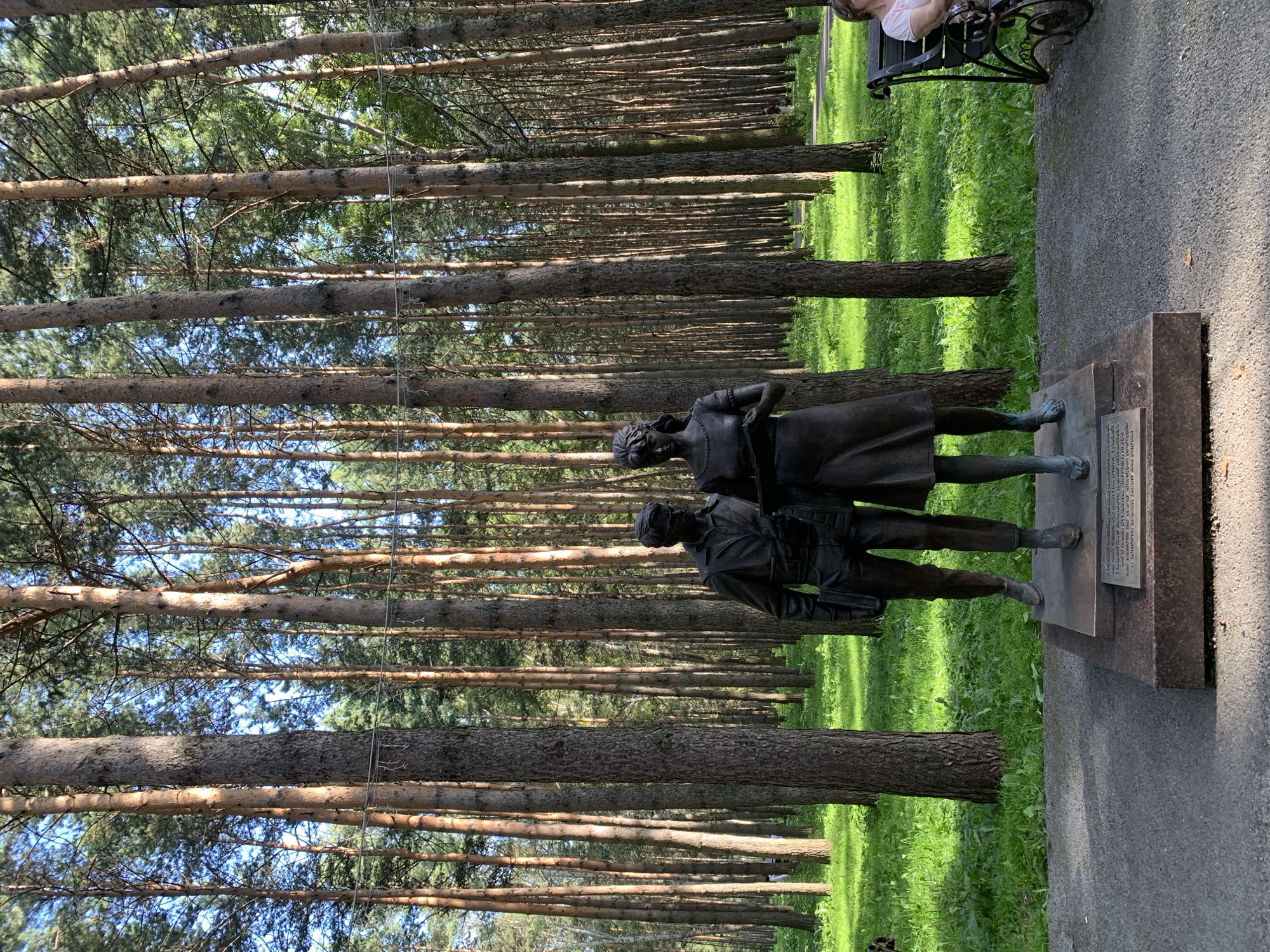 This is a full, eye-level, medium outdoor shot of a bronze statue of a man and a woman standing side-by-side in a park. The statue is on a granite pedestal. The man is on the left, wearing a jacket and trousers, and holding a book. The woman is on the right, wearing a dress and holding a book. The statue is in a park with tall pine trees behind it. The ground is covered in grass and gravel. In the background, to the right, a woman is sitting on a park bench. The statue is likely located in Prokopyevsk, Russia.