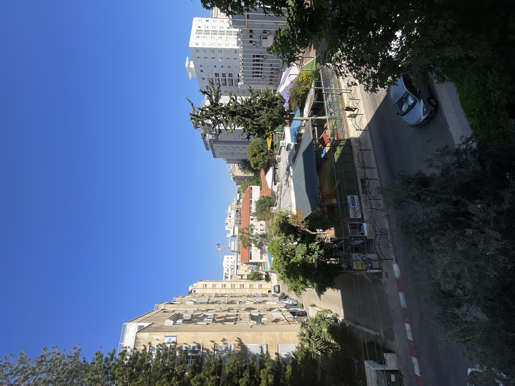 The image shows a residential street in Netanya, Israel, on a clear day. The view is from a slightly elevated position, looking down onto a street lined with apartment buildings and trees. On the left side of the street, a tan-colored apartment building with multiple windows and air conditioning units is visible. Several cars are parked along the narrow street in front of the building.
Across the street, there is a playground area with colorful equipment, including slides and climbing structures, enclosed by a fence. Several small buildings with awnings are part of the playground or adjacent facilities. Beyond the playground, more apartment buildings of varying heights and architectural styles are visible, including a modern white building with many windows and a light blue building with a distinctive facade.
A single silver car is parked on the right side of the street, partially shaded by trees. Two individuals are walking on the sidewalk near the playground area. The sky is a clear, deep blue, suggesting a sunny day. The overall scene depicts a typical urban residential environment in Netanya.