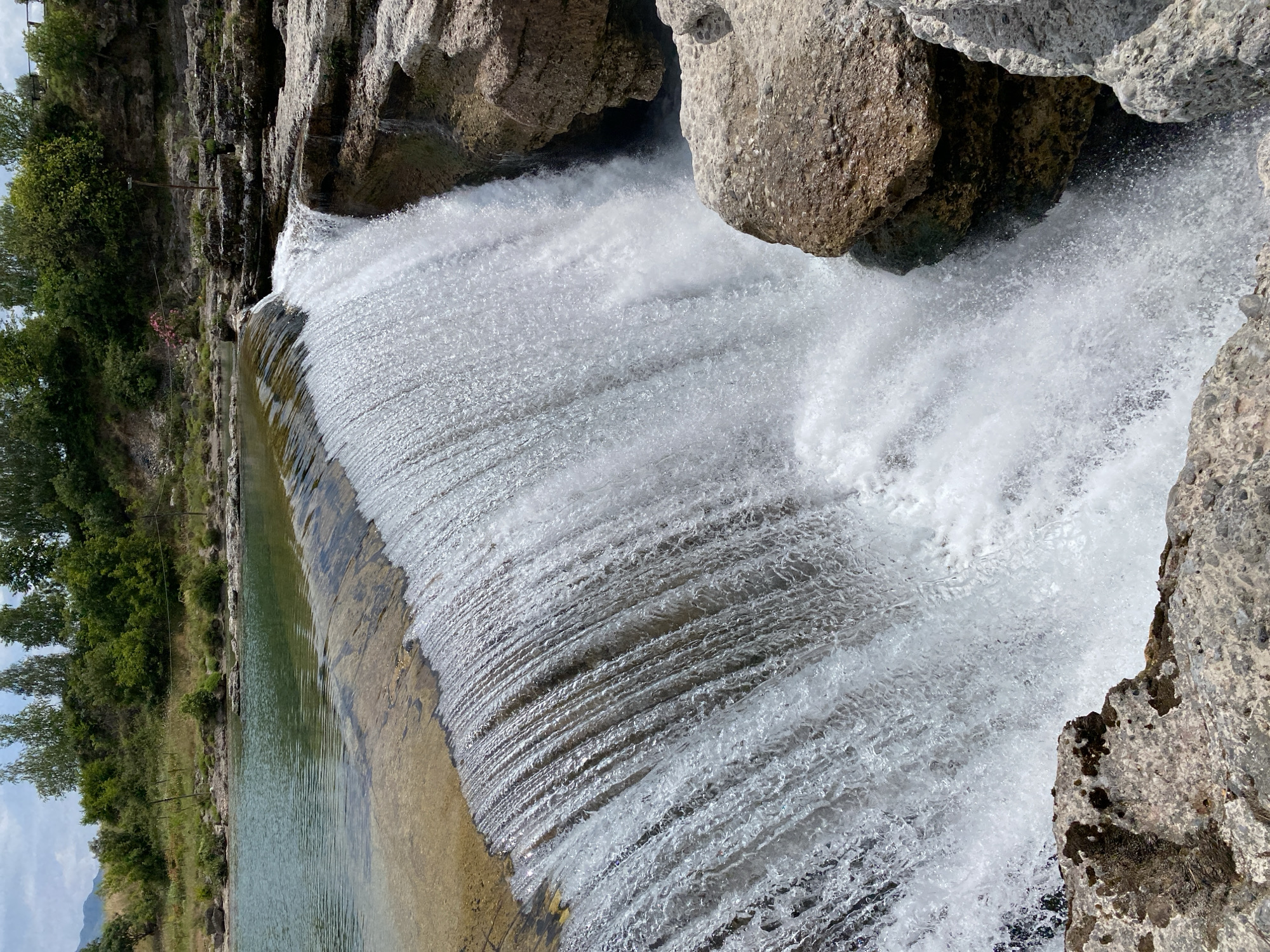 The image displays a wide, natural waterfall or cascade, likely the Cijevna Falls near Podgorica, Montenegro, captured under bright daylight. Water flows powerfully over a series of relatively flat, textured rock formations, creating a broad, frothy white curtain as it cascades downwards. Above the main drop, a serene body of clear water reveals the rocky riverbed below, reflecting the sky.
The dynamic waterfall descends into a turbulent pool, generating significant spray and foam. Large, rugged rocks, varying in shades of grey and brown and showing distinct textures, frame the waterfall and define its banks. On the left side of the scene, lush green trees and dense bushes line the riverbank, extending into the background, with distant rocky hills visible under a light blue sky dotted with white clouds. Faint man-made elements, possibly a wooden post and wires, are discernible among the vegetation.
No people or visible text are present in the image. The scene conveys the raw beauty and powerful flow of nature on a clear, sunny day.