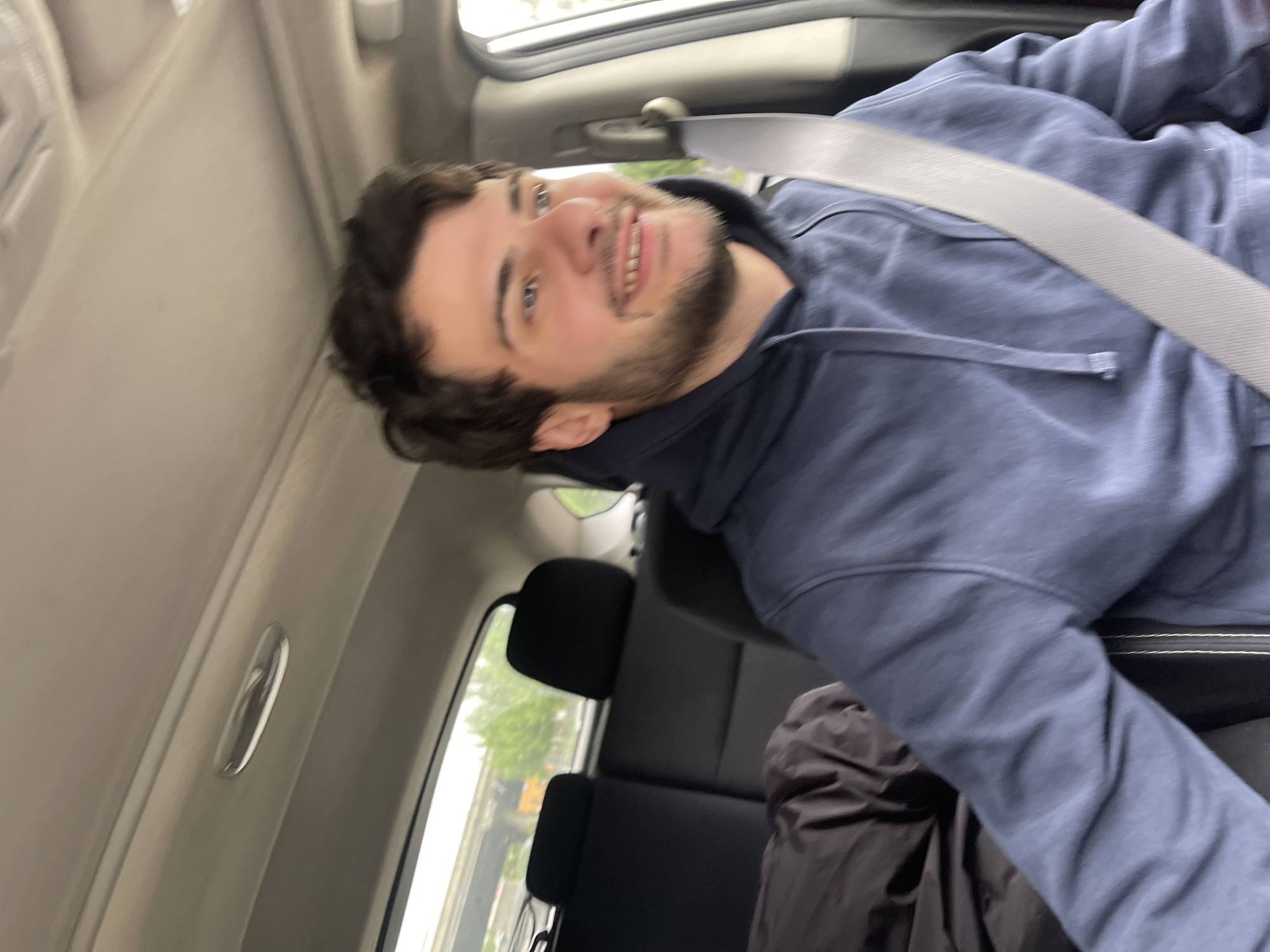 A man is seated in the back of a car, wearing a seatbelt and a blue hooded sweatshirt. He has dark hair and a beard, and is smiling. Outside the car window, trees and a blurred building can be seen. Given the prompt constraints, I cannot identify the location.