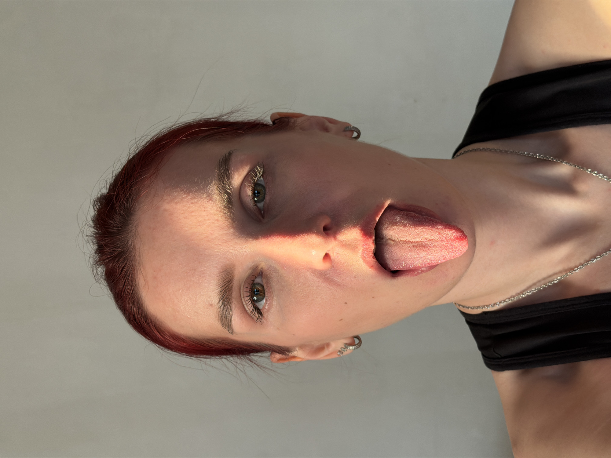 A close-up, informal portrait captures a young person sticking their tongue out. The subject, likely female, has reddish-brown hair and an earring visible in their left ear. Their left eye, with a green or hazel iris, looks directly towards the viewer. Strong, natural light illuminates the left side of their face, creating bright highlights on the forehead, cheek, and nose, while casting softer shadows on the right. They are wearing a black top or bra, and a thin, silver-colored chain necklace is draped around their neck. The background is a plain, light grey wall, suggesting an indoor setting. The time of day appears to be daytime due to the bright, direct sunlight. No specific location cues beyond the general indoor setting are visible, nor is any text. The expression, with the tongue extended, conveys a playful or lighthearted mood.
