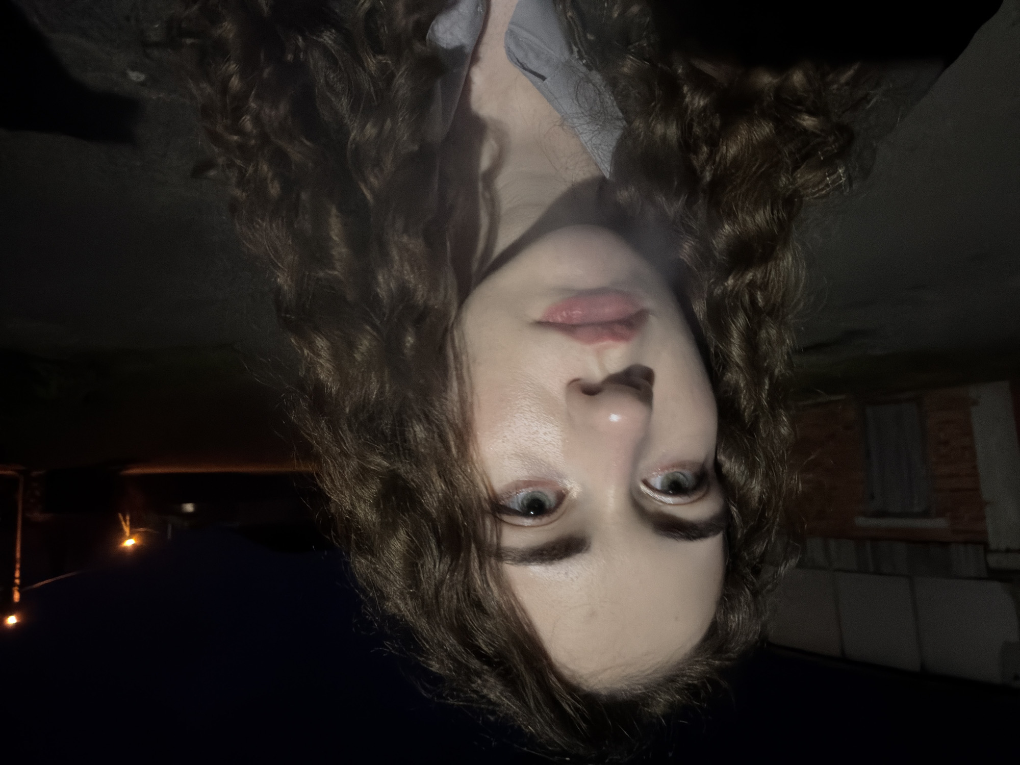 A young woman with curly brown hair is pictured in a close-up, low-light, outdoor scene. She is looking slightly to the side, her face illuminated by a nearby light source. Her expression is neutral. In the background, a brick building with a large door is visible on the left, and two streetlights can be seen in the distance on the right. The ground appears to be asphalt. The setting is likely an urban or suburban area within Zavodoukovsk, Russia, given the presence of streetlights and a building.