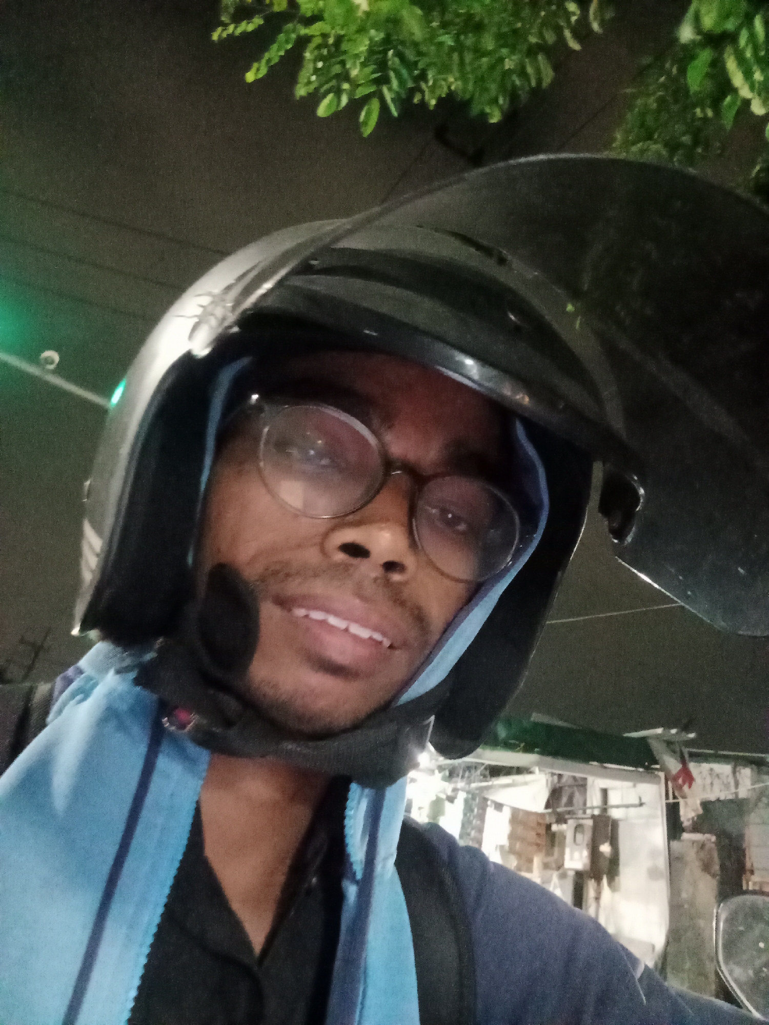 A close-up, low-light, selfie-style photo shows a person wearing a black helmet and round glasses, with their head turned towards the camera. The person appears to be smiling slightly, revealing their teeth. They are wearing a blue scarf or hood around their neck and head, and a dark shirt underneath. In the background, there are blurred elements of what appears to be a street scene at night, with some foliage visible at the top of the frame. The image seems to have been taken from a moving vehicle, likely a motorcycle, given the helmet. The overall lighting suggests it is nighttime or dusk. There are no specific visual cues that definitively place the location in Surabaya, Indonesia, beyond the provided information.