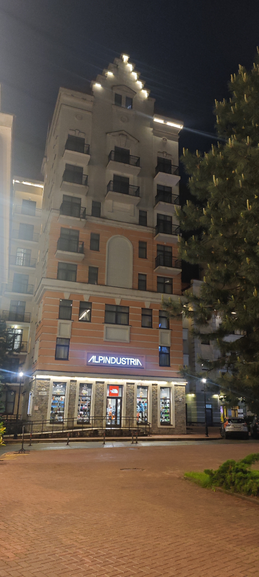 A nighttime view of the Alpinindustria store in Estosadok, Russia. The store occupies the ground floor of a multi-story building with a distinctive architectural style, including balconies and a stepped gable roof. The store's illuminated sign is clearly visible above the storefront. A tree is seen in the right foreground. The ground is paved with red brick. A car is visible parked nearby.