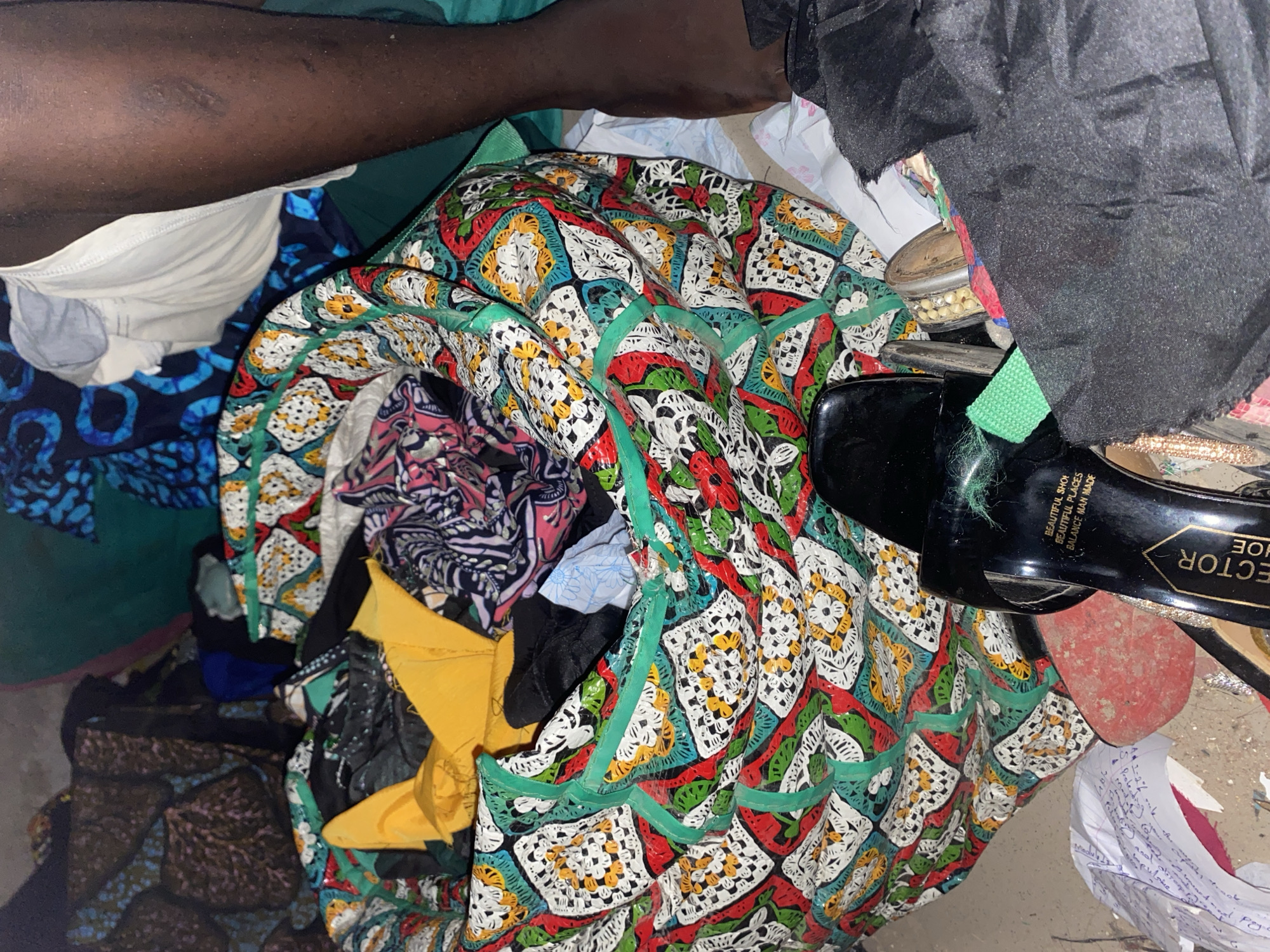In Jauro Yinu, Nigeria, the image depicts a chaotic scene of scattered textiles and personal belongings, suggesting a state of disarray or transition. A large, colorful patterned bag, possibly a laundry bag, is centrally placed and overflowing with clothes. Among the items are yellow fabric pieces, black clothing, and patterned garments, creating a vibrant yet cluttered visual. A black sandal with white text is visible in the foreground, with part of its sole peeking out. Nearby, a woman's dark-skinned leg is visible, extending from the upper right corner of the frame, with a portion of white undergarments also present. The environment appears to be indoors, given the presence of various fabrics and the lighting, though the floor surface is not clearly discernible. The overall impression is one of a lived-in space where items have been hastily placed or are in the process of being organized.