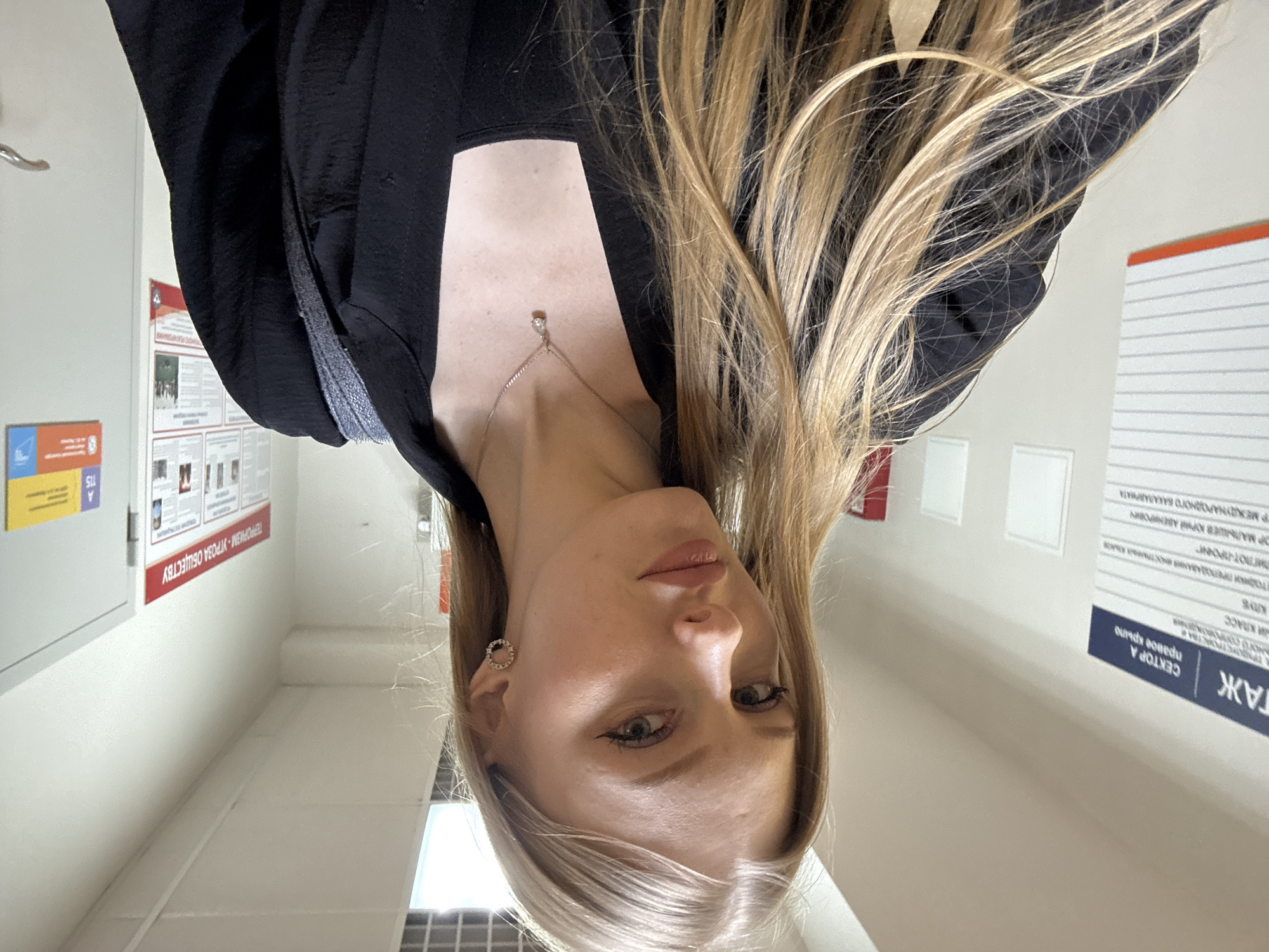 A woman with blonde hair is photographed indoors, in what appears to be a corridor or hallway with signage in Russian. She is wearing a black blazer or top, and a delicate necklace with a pendant. The signage visible in the background includes text related to education, sector A (right wing), teaching methods and a warning about terrorism. The location may be Perm, Russia, based on the Cyrillic script in the background and the image parameters.