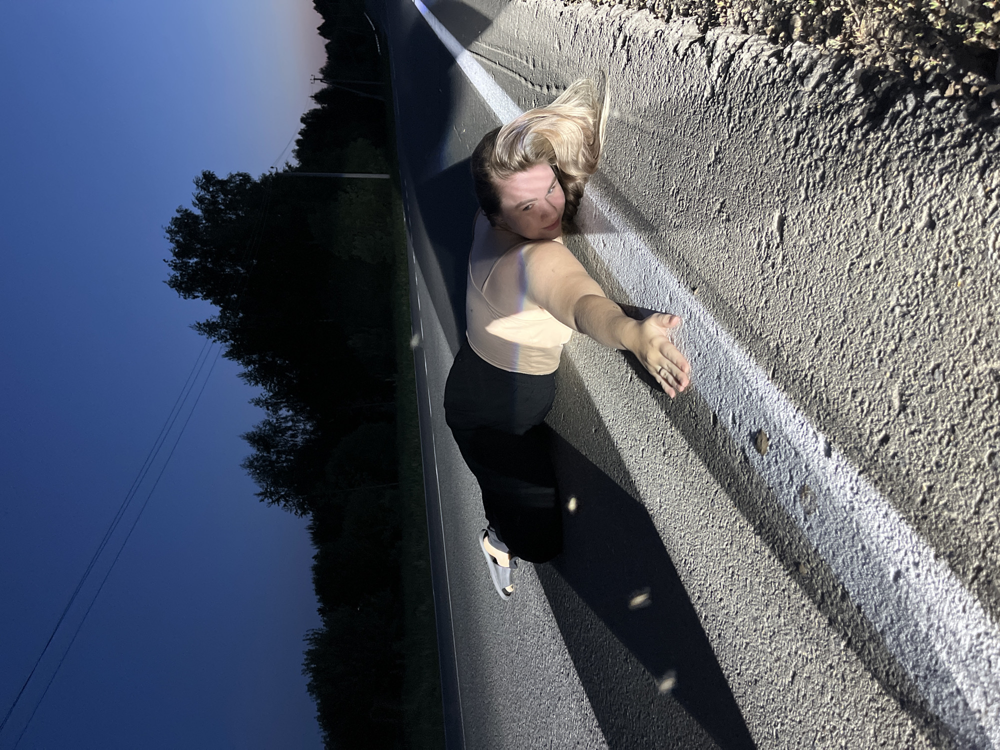 A woman is standing on the shoulder of a road in Olkhovka, Russia. The road has a solid white line marking the edge of the lane. The woman is wearing a light-colored top, dark pants, and sandals. She has blonde hair and is looking over her shoulder at the camera. Her right arm is extended, with her hand near the white line. The background consists of trees silhouetted against a dark blue sky, suggesting it is dusk or night.