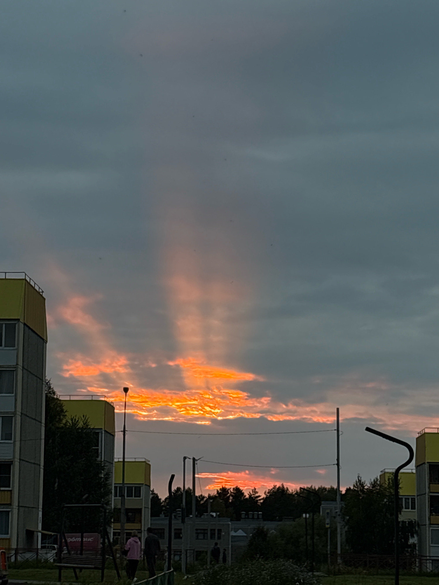 The image depicts a sky filled with dramatic crepuscular rays, also known as "sunbeams," emanating from a bright, fiery sunset. The sunbeams are cutting through a grey, overcast sky, illuminating scattered clouds with vibrant orange and pink hues. In the foreground, residential buildings with yellow and grey facades are visible, suggesting an urban or suburban setting. The architecture appears to be functional, characteristic of Soviet-era apartment blocks. Trees and other vegetation are present, adding a touch of nature to the built environment. A street lamp stands prominently in the mid-ground, its dark silhouette contrasting with the bright sky. A swing set and a bench are visible in the lower left quadrant, indicating a playground area. A few individuals are present in the scene, walking or standing, their figures small against the backdrop of the sky and buildings. The overall impression is that of a picturesque sunset over a residential area, possibly in a northern European climate given the overcast sky and architecture. The location is identified as Марковский, Russia.