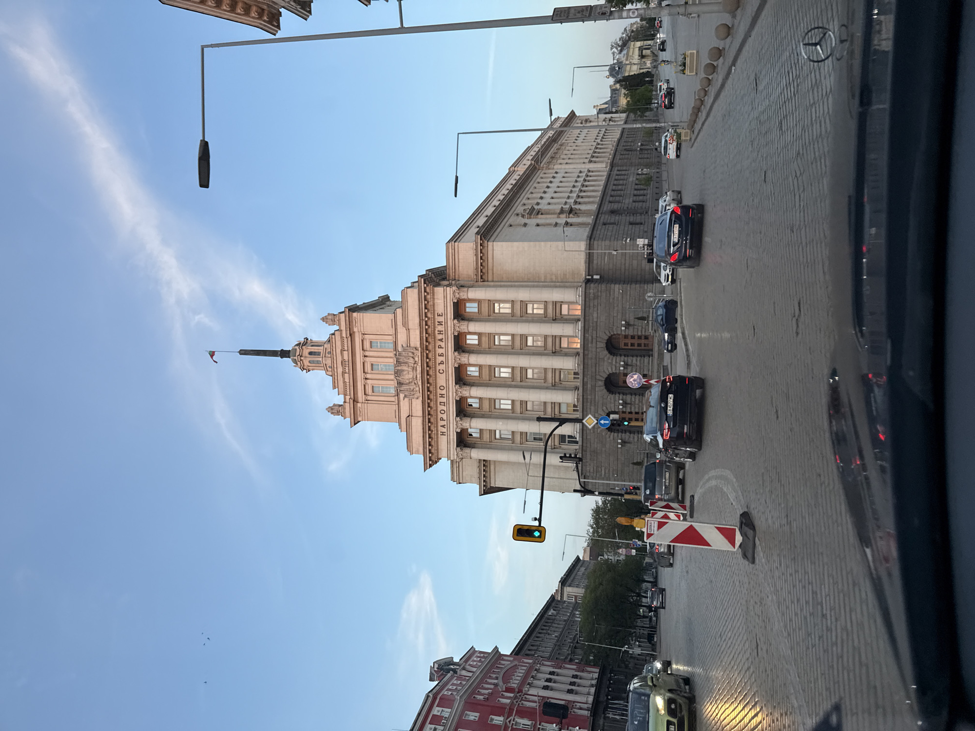 The image, when viewed upright, captures a vibrant urban street scene in Sofia, Bulgaria, centered around the majestic National Assembly building. This prominent structure, on the left, showcases classical architecture with light-colored stone, numerous columns, and a distinguished multi-tiered spire topped with a small flag. The building's name, "НАРОДНО СЪБРАНИЕ" (National Assembly in Bulgarian), is clearly visible on its facade.
To the right, a wide cobblestone street is bustling with cars, indicating active traffic. Streetlights with sleek, modern designs are interspersed along the road, alongside functional traffic lights—one displaying yellow, another green—and various blue directional road signs. In the immediate foreground on the street, red and white striped construction barriers are noticeable. The perspective suggests the photograph was taken from inside a vehicle, with a portion of a dashboard or window frame featuring a Mercedes-Benz logo visible on the far right.
The sky above is a clear blue, adorned with subtle, wispy clouds, suggesting a time of day in the late afternoon or early evening, given the quality of natural light and the illumination of some car headlights. In the bottom left corner, a section of a red building with white window frames adds a touch of color to the scene. The overall impression is one of an active yet orderly European city street.