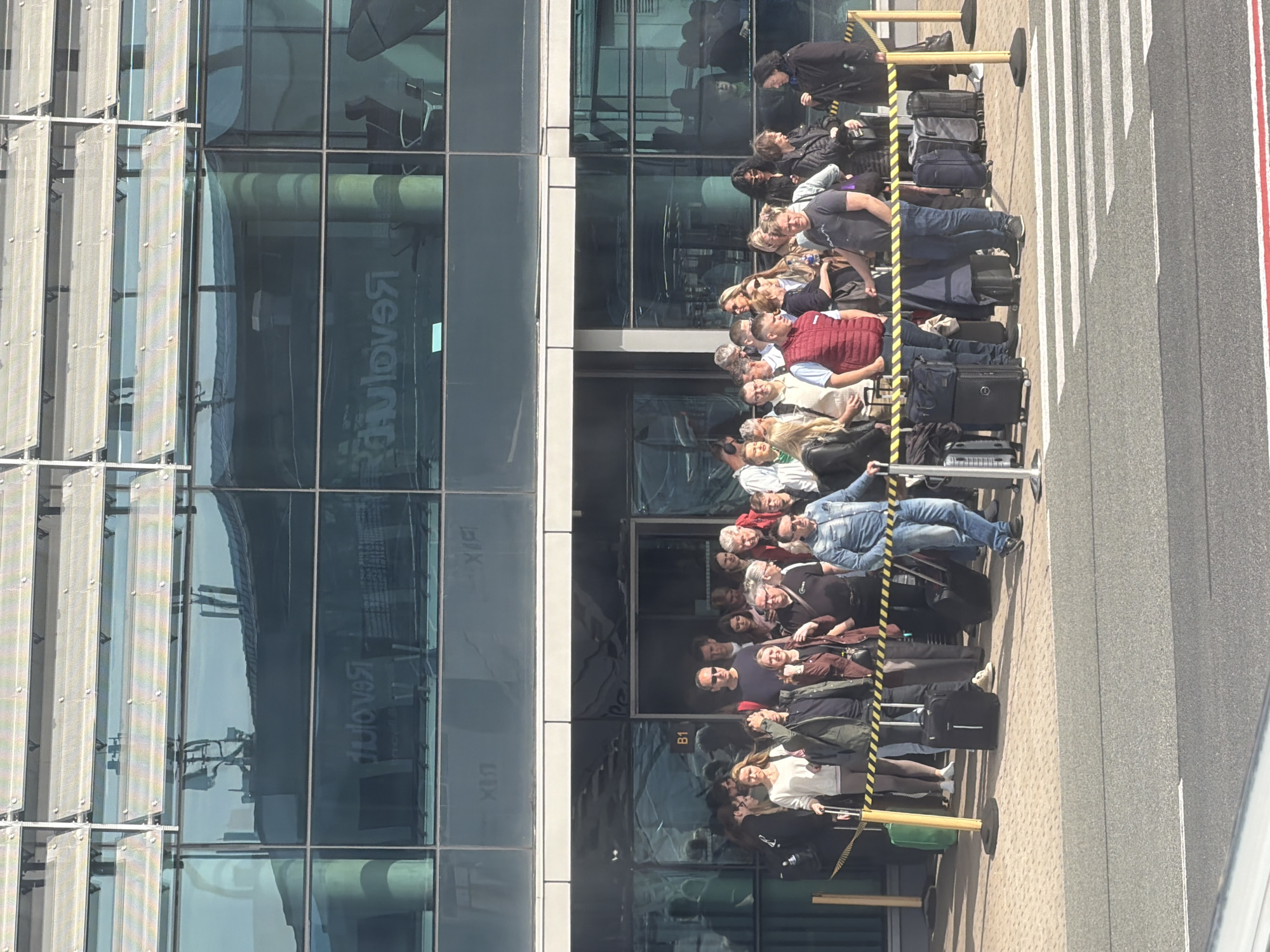 The image shows a large group of people standing in a line outside a modern building with a glass facade. The people are diverse in age and appearance, and most are holding luggage. The building has reflections of other structures in its windows. The line is delineated by yellow and black stanchions. The ground next to the line is paved with a crosswalk pattern. The image is taken outdoors in daylight. The location could be an airport or transport hub based on the presence of travelers and luggage. It is unclear whether this image relates to Skulte, Latvia. Further information is needed to confirm this.