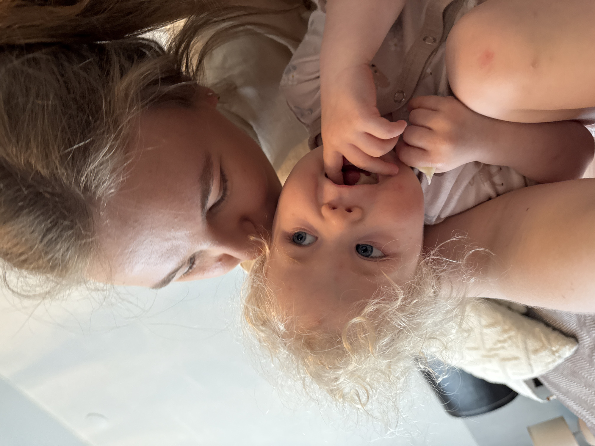 An intimate, low-angle portrait captures an adult and a young child in a tender interaction, set indoors in Vladimir, Russia. The image is taken from a perspective looking up at the subjects. On the left, an adult, likely female, with light brown hair, leans in close to the child, her eyes gently closed or looking downwards, a soft smile visible.
The young child, situated towards the center-right, has bright blue eyes and a mass of curly blonde hair, looking directly towards the camera with fingers playfully inserted into their mouth. The child wears a light-colored, button-up outfit, and a small red mark is visible on their right elbow. Their bodies are in close proximity, suggesting a cuddle or playtime.
The background is brightly lit and softly blurred, indicating a casual indoor setting, possibly a home, with a glimpse of a patterned, light-colored pillow or blanket near the child's head. The overall atmosphere conveys warmth, affection, and joy. No text is visible in the image.