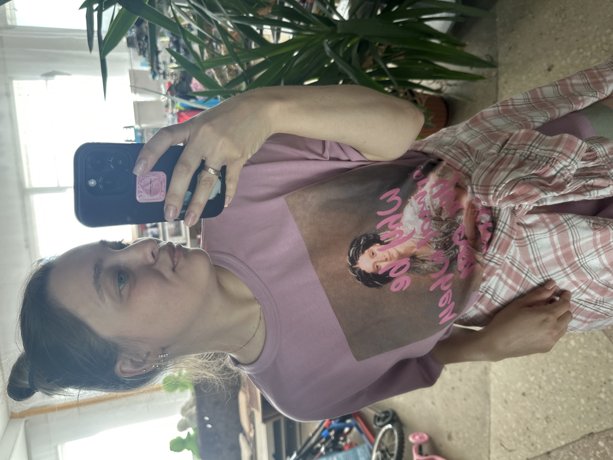The image shows a young woman taking a mirror selfie in what appears to be a residential setting, potentially within Yaroslavka, Russia. She is wearing a pink t-shirt with a portrait of a woman on it and a pink and white plaid shirt tied around her waist. She is holding a smartphone in her right hand, and a ring is visible on her left ring finger. The background includes a large potted plant, furniture, and windows suggesting an indoor space. The lighting is natural, coming from the windows.