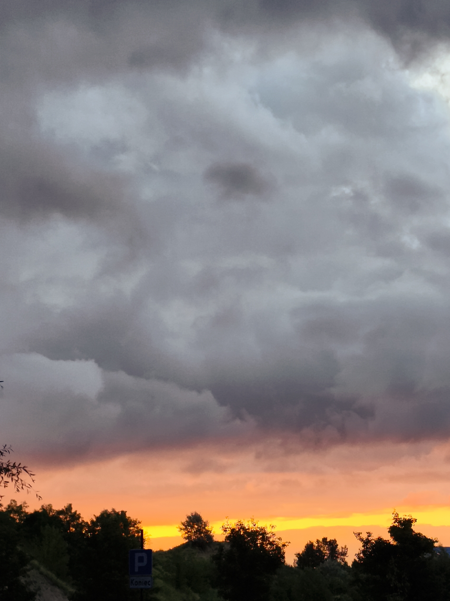 This is a photograph of a dramatic sunset over Gdańsk, Poland. The sky is filled with dark, heavy clouds that are tinged with shades of gray and purple. Below the clouds, a vibrant band of orange and yellow light stretches across the horizon, indicating the setting sun. Silhouetted against the colorful sky are the dark shapes of trees and vegetation. In the foreground, a blue parking sign with the letter "P" and the Polish word "Koniec" (which means "End") is visible. The scene evokes a sense of impending weather change or the end of a day.