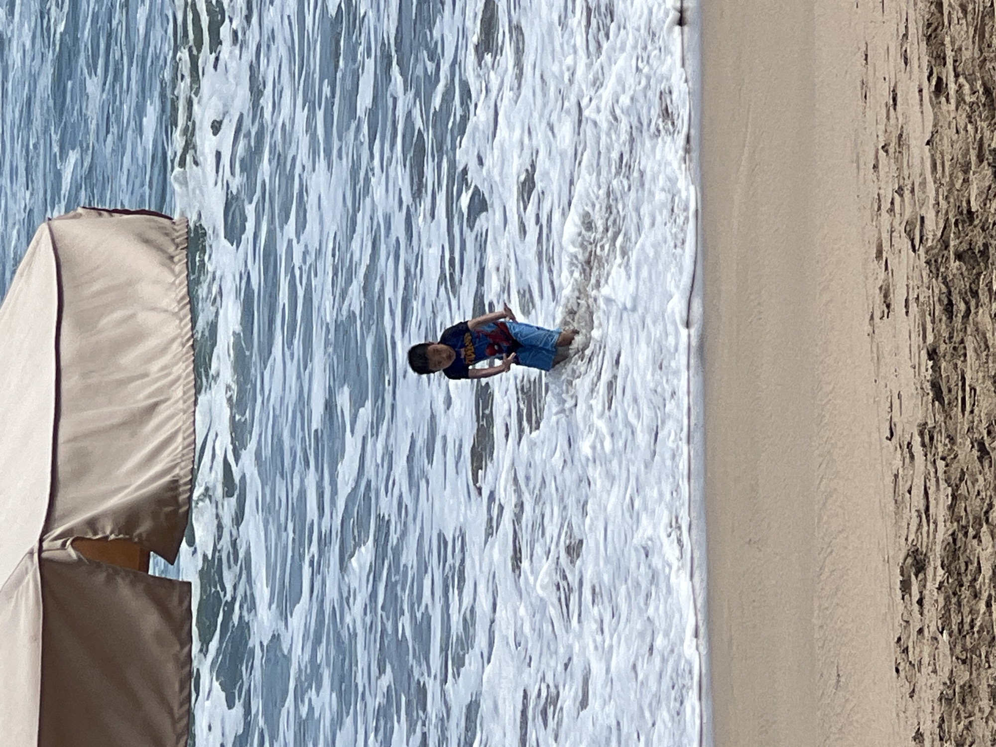 A young boy with dark hair is standing in the shallow water near the shore at Kuta Beach, Bali, Indonesia. He is wearing a dark t-shirt with a graphic design and blue shorts. The boy is facing towards the camera and appears to be wading or playing in the foamy waves. The ocean water is blue with white foam from the waves, and the beach sand in the foreground is light brown with some debris. In the background, a large, light-colored canopy or umbrella is partially visible, extending over the water. The overall scene depicts a typical beach day with a child enjoying the ocean.