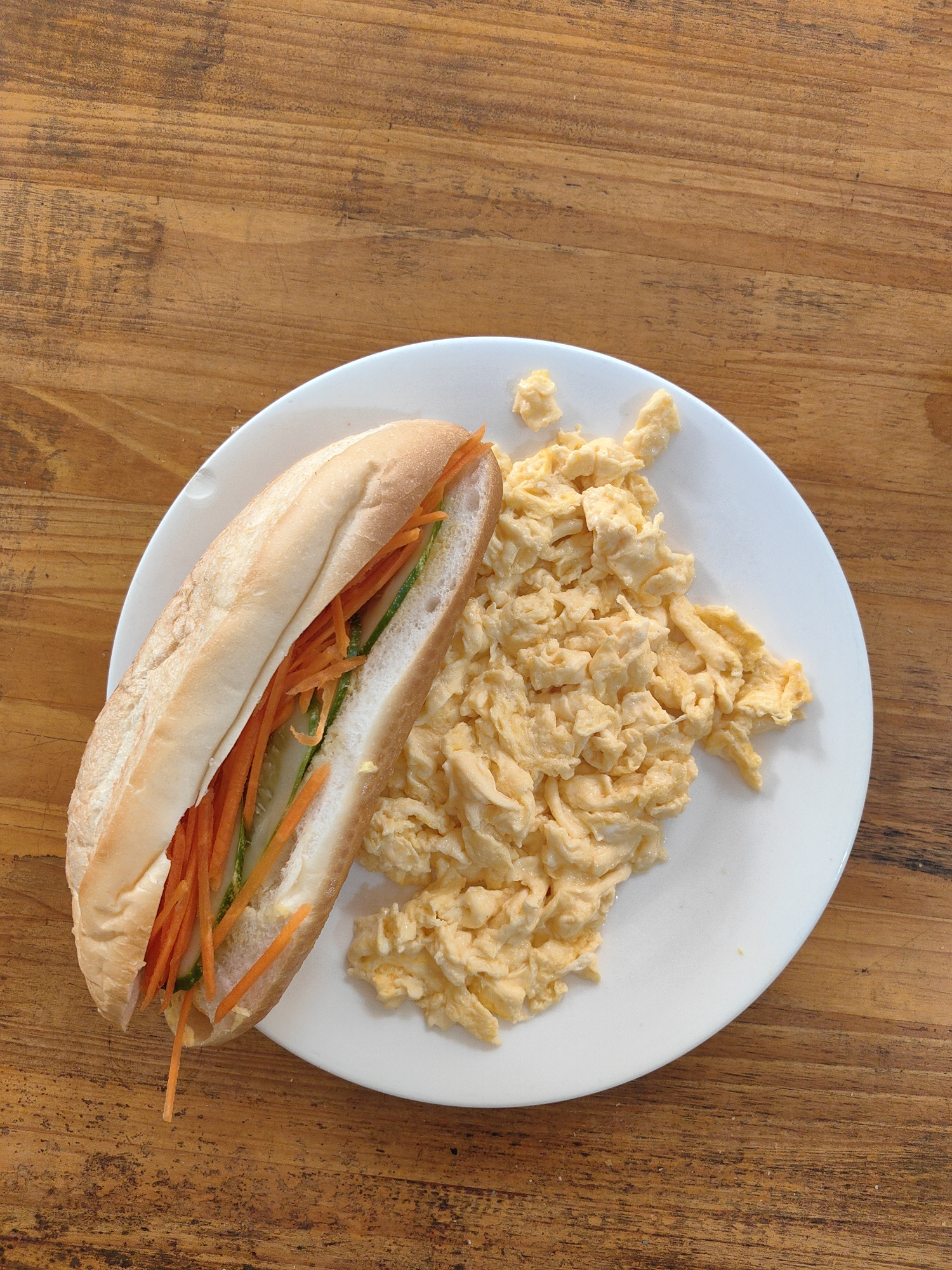 A top-down view of a white plate containing a Vietnamese bánh mì sandwich and scrambled eggs, placed on a wooden table. The bánh mì is filled with julienned carrots and possibly other pickled vegetables. The scrambled eggs are yellow and appear soft. The wooden table has a warm, brown hue and visible grain. The plate and food appear indoors under natural lighting. The image does not provide information to confirm its location in Bai Chay Ward, Vietnam.