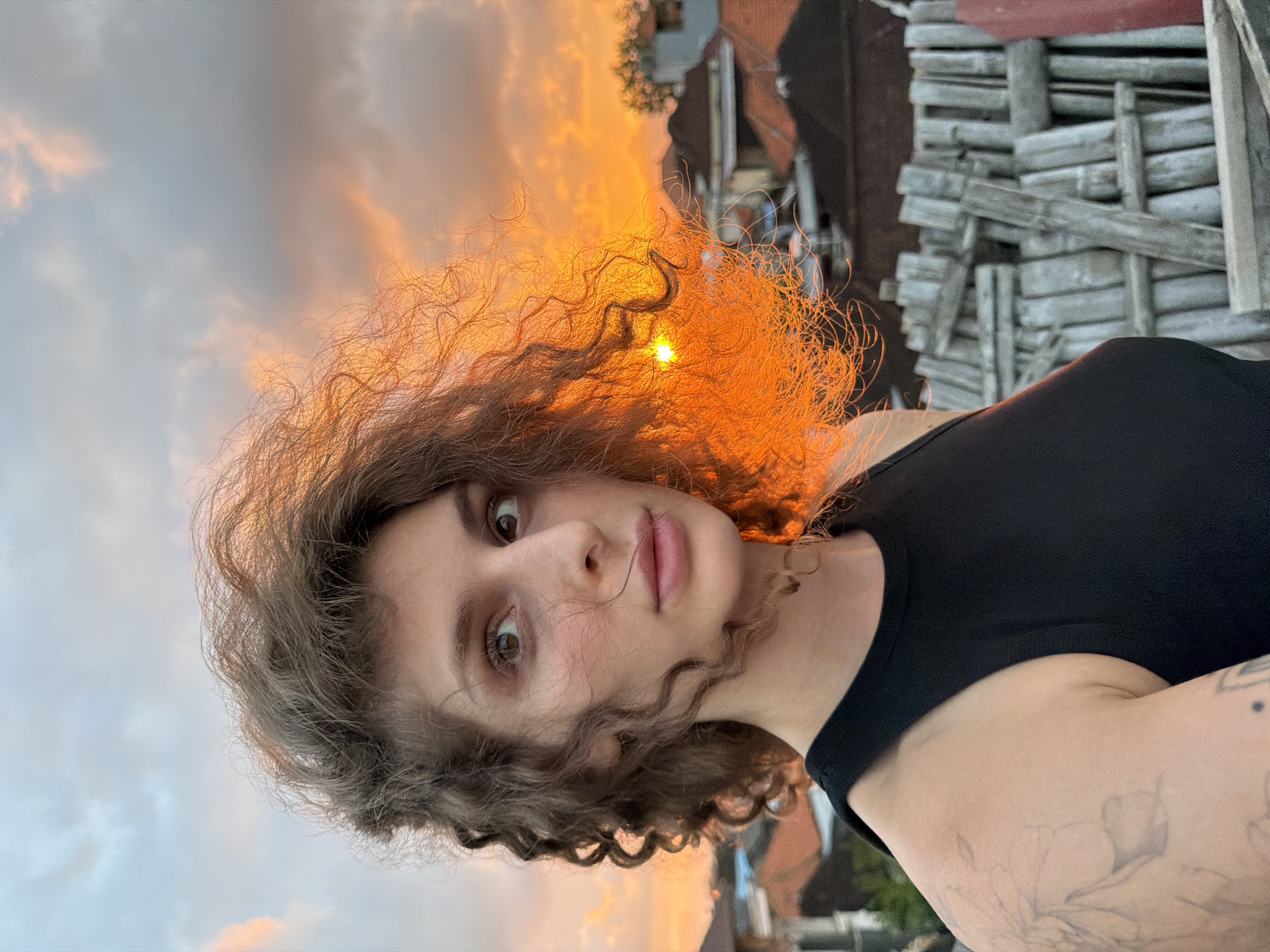 A person with curly brown hair is positioned in the foreground, looking to the left of the frame. Their hair is illuminated by the setting sun, creating a bright orange glow around their head. They are wearing a black tank top. The background shows a cityscape with red-tiled roofs, and a structure made of weathered wooden poles or bamboo. The sky above is cloudy with hues of orange, pink, and blue, indicative of sunset. The location is identified as North Kuta, Indonesia.