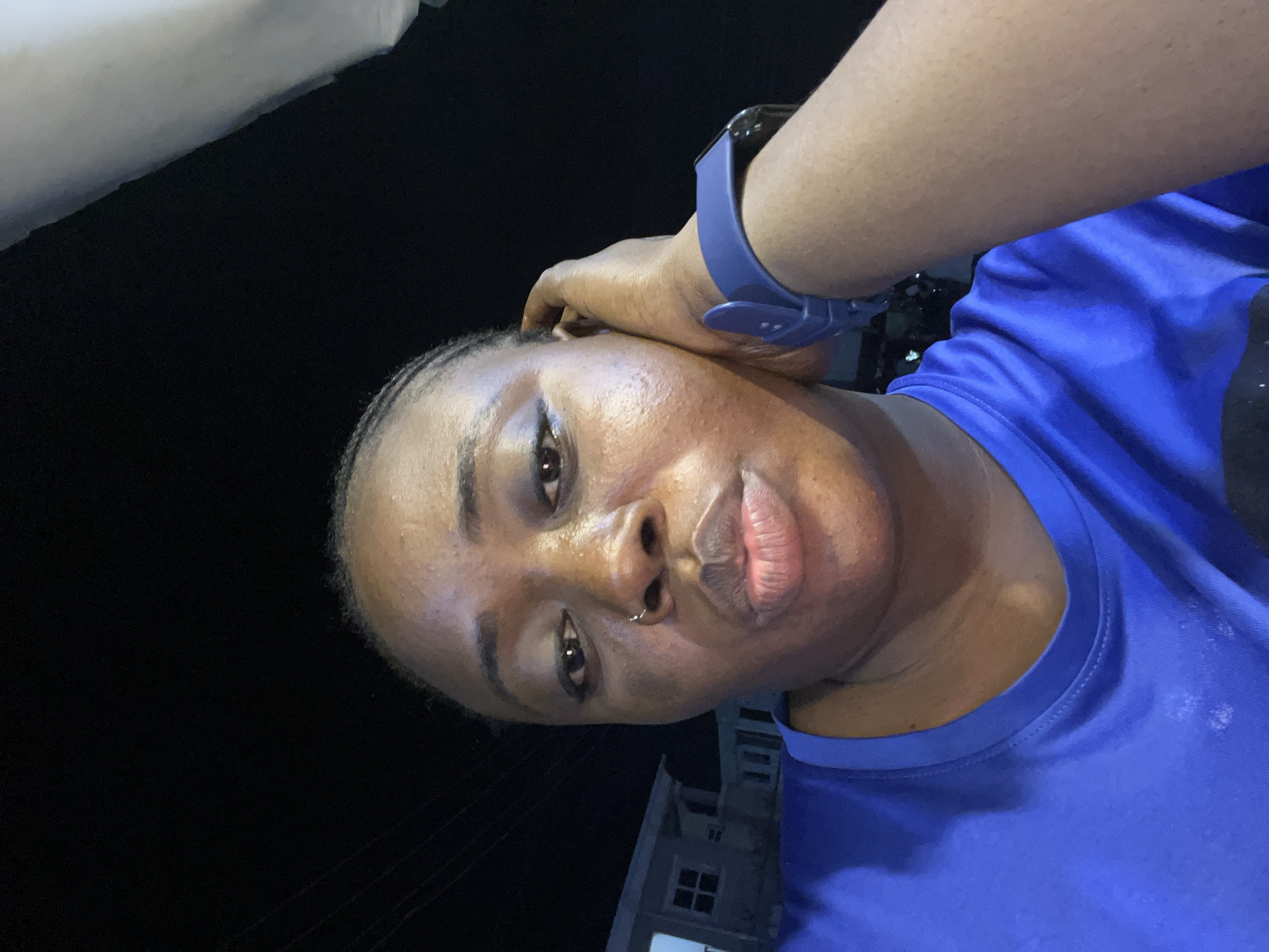A close-up, selfie-style photograph shows a dark-skinned individual with their hand on their head. The individual wears a blue sports watch and a blue t-shirt. The background is dark, revealing a building with a window, partially illuminated. The location is Kubwa, Nigeria.