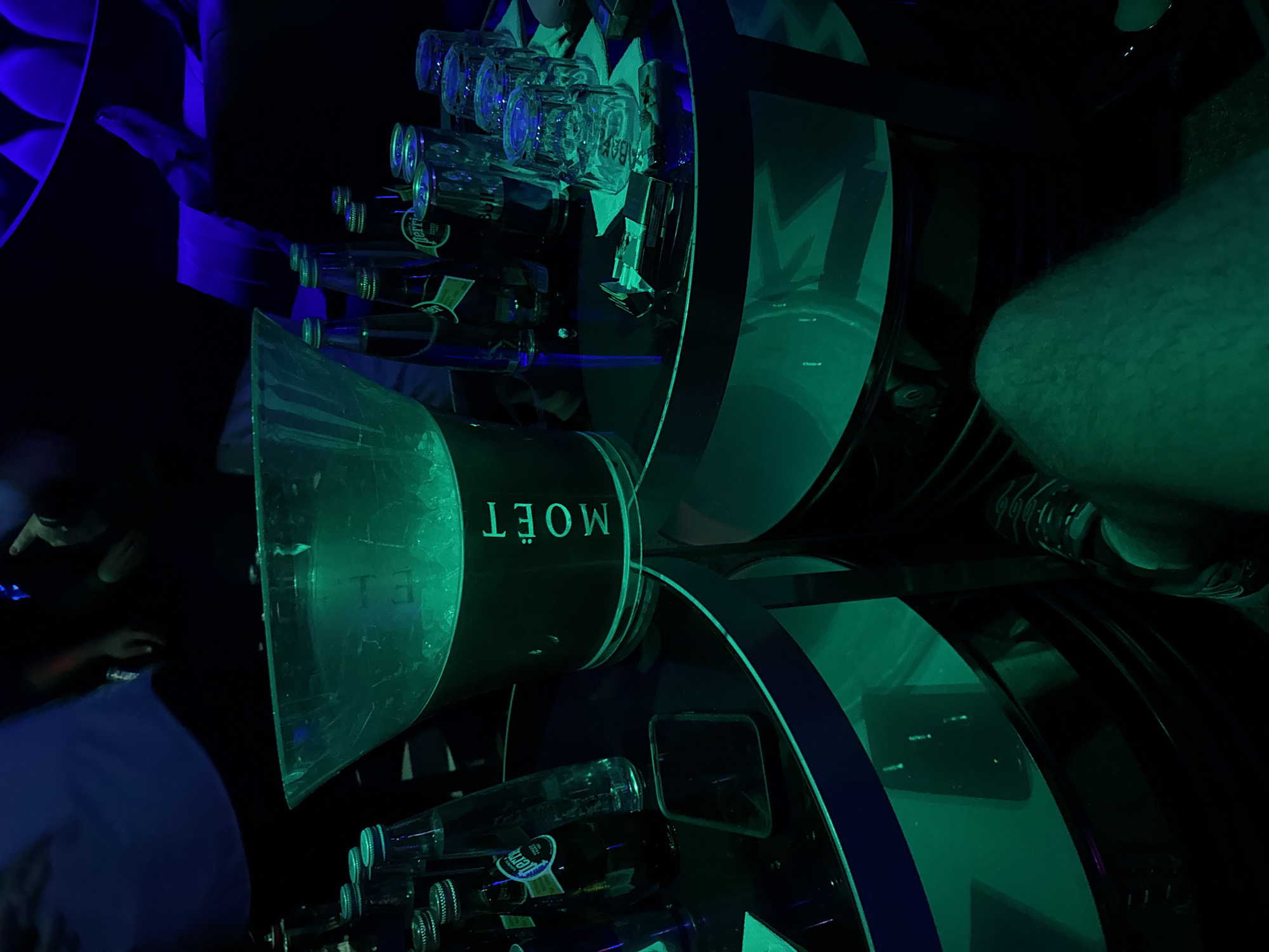 The image shows a circular table at a club in Plovdiv, Bulgaria. The table is dark and illuminated by green and blue lighting. On the table are several bottles of alcoholic beverages and empty glasses. There is a Moët & Chandon ice bucket. The table appears to be at a club, as inferred by the lighting conditions and drinks. People are partially visible on the periphery of the table.