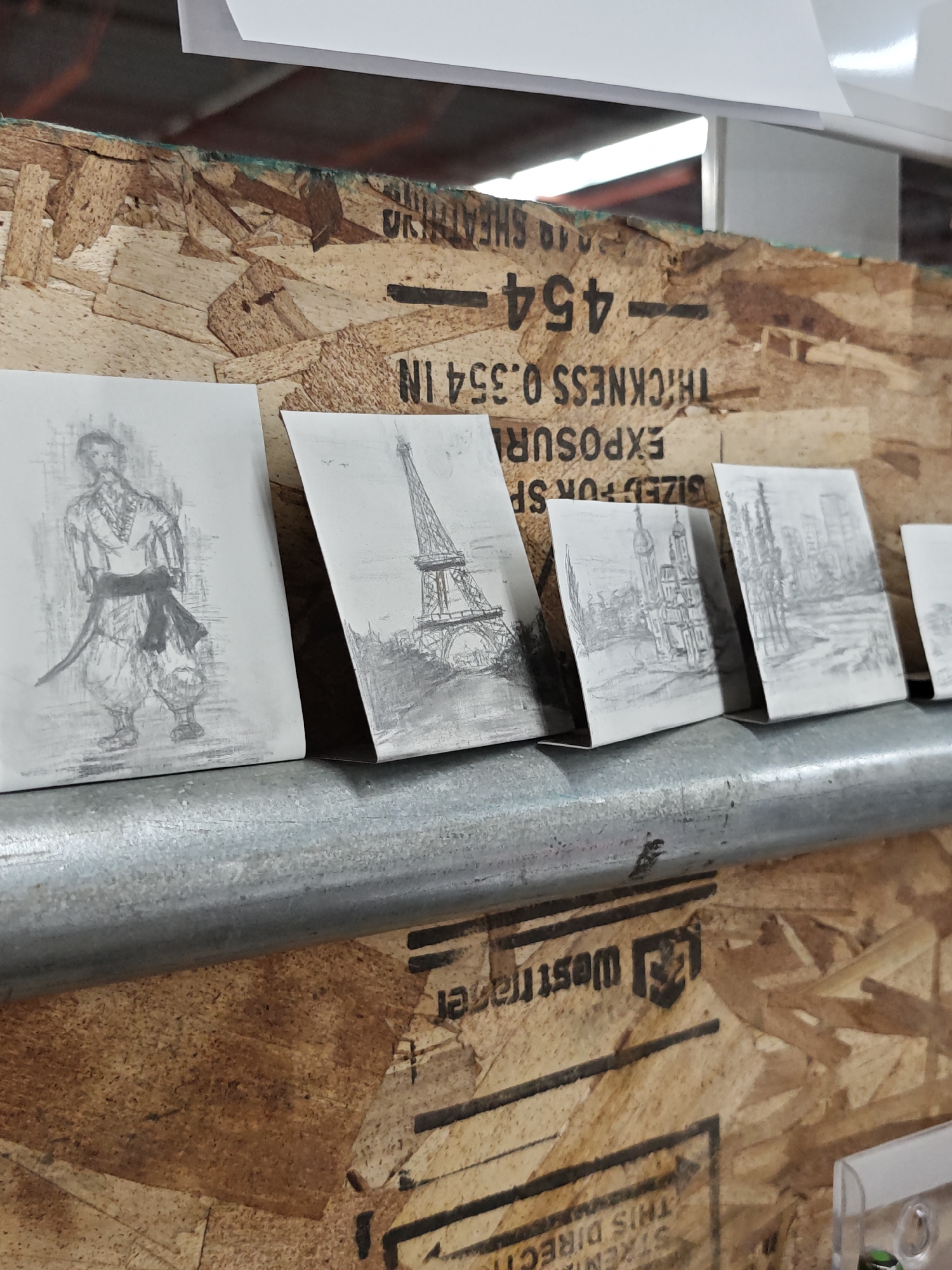 The image shows a shelf displaying several small pencil sketches. The sketches are of various subjects, including a samurai warrior, the Eiffel Tower, and cityscapes. They are placed on what appears to be a piece of plywood, which is mounted on a metal pipe. The background is a wooden surface with some text printed on it, including "WESTERN" and "EXPOSURE". The scene appears to be indoors, possibly in a workshop or art studio. There is no indication that this scene is located in Calgary, Canada.
