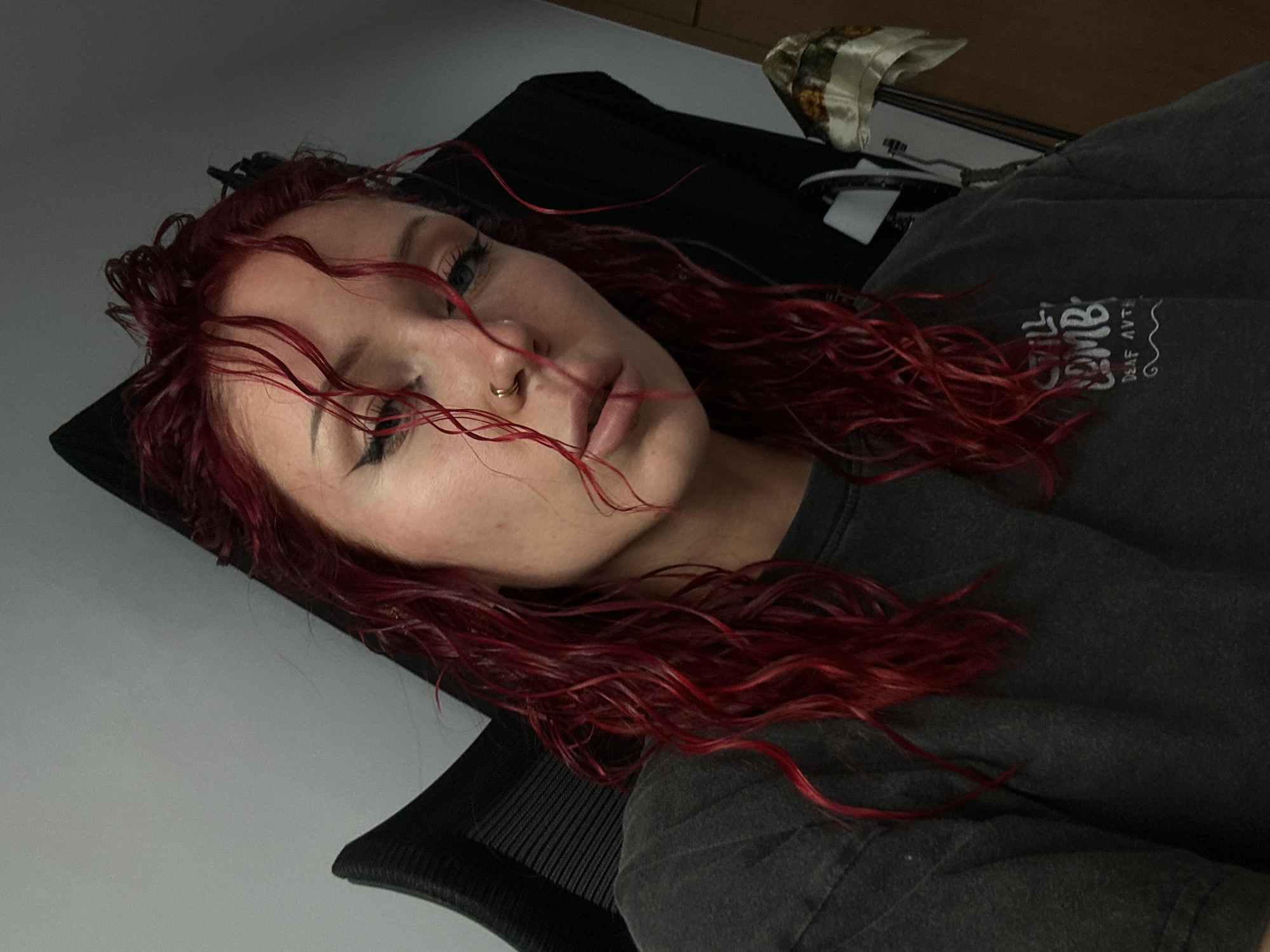 A woman with red, wavy hair lies reclined in a black office chair. She has a nose ring and dark eyeliner, and she is wearing a dark gray t-shirt with white text. The background includes a gray wall and a wooden cabinet with a patterned cloth on top. The image was taken indoors. There is no evidence in the image to suggest it was taken in North Kuta, Indonesia.