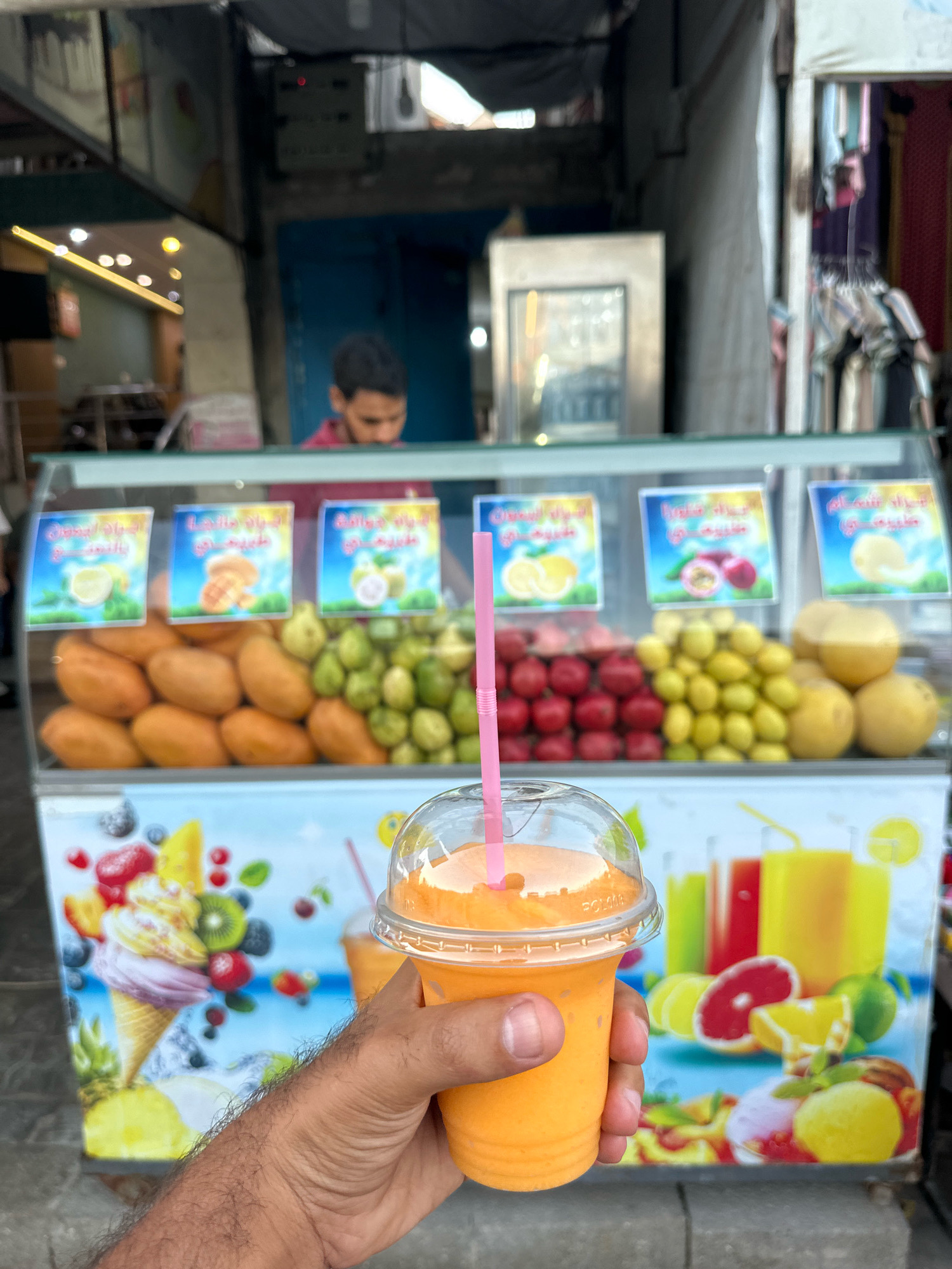 A person's hairy arm holds a clear plastic cup filled with a vibrant orange beverage, topped with a clear plastic lid and a pink straw. The cup is positioned in the foreground, obscuring the lower half of the image. In the background, a fruit stand displays an assortment of fresh fruits, including mangoes, guavas, and pomegranates, arranged behind a glass counter. Signs with Arabic script and illustrations of fruits are visible above the fruit displays. A man wearing a pink shirt is partially visible behind the counter, attending to the stand. The stand itself is adorned with a colorful graphic depicting ice cream cones and various fruits. The scene appears to be an outdoor market or street vendor stall in Gaza City, Palestinian Territories, with a backdrop suggesting a bustling urban environment. The overall lighting is bright, indicating daylight.