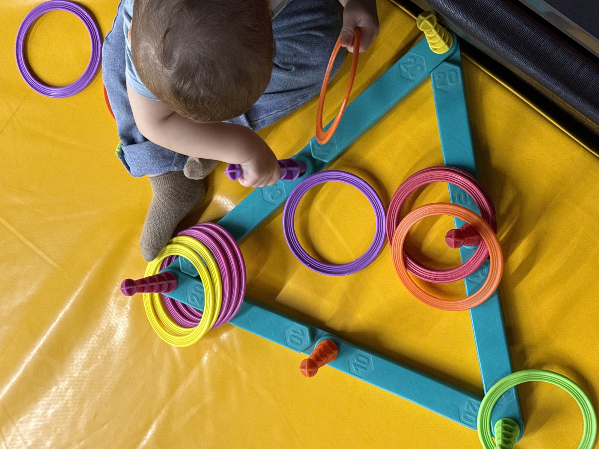 The image displays a close-up, overhead view of a baby playing with a ring toss toy on a yellow mat. The ring toss toy is teal and triangular in shape, with several pegs. Rings of various colors—yellow, purple, pink, orange, and green—are scattered around the toy or stacked on its pegs. One of the baby's hands is visible, holding a purple ring. The baby is wearing a light blue shirt and denim overalls. The background is a textured yellow surface, possibly a mat or tarp. The overall scene is brightly lit, suggesting an indoor setting. There is no information available to determine the location in Krasnoyarsk, Russia, or any other specific geographical context.