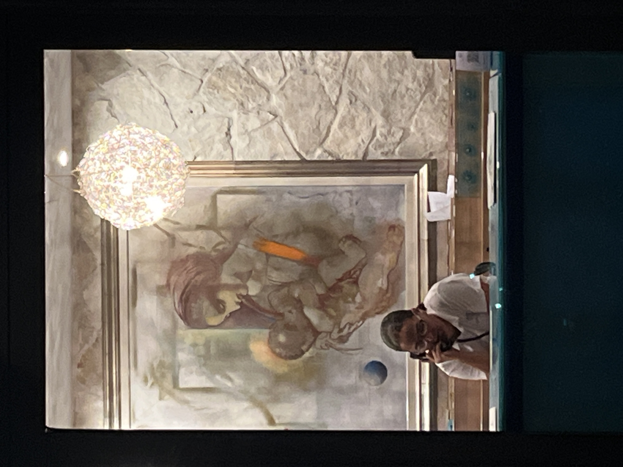 A woman wearing glasses and a white shirt is sitting at a desk and talking on a black corded phone. She is looking down and her right hand is holding the receiver to her ear, while her left hand is resting on the desk. Behind her, a large framed painting depicts a woman holding a baby. The painting is set against a wall with a stone texture. A decorative light fixture, resembling a spherical cluster of lights, hangs from the ceiling to the right of the painting. The desk has various items on it, including what appears to be a small blue sphere in the painting, a crumpled piece of white paper, and other office supplies. The overall scene appears to be an indoor setting, possibly a reception area or office, with the dark foreground suggesting the viewer is looking through a window or doorway. The location is identified as Budva, Montenegro.