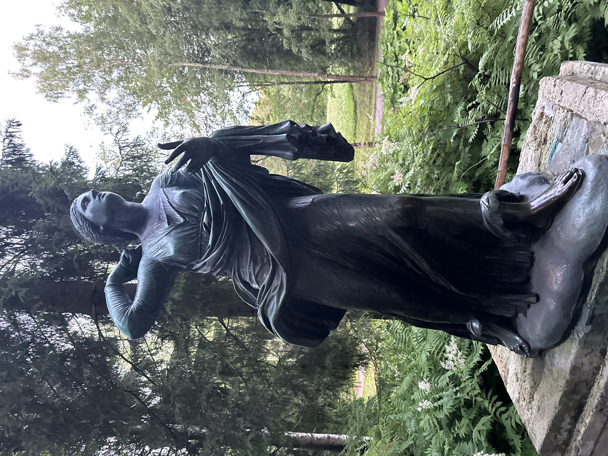 The image shows a full-body bronze sculpture of a draped female figure standing on a stone pedestal. The sculpture depicts the figure with her left arm raised, holding a piece of fabric, and her right arm extended forward, with her fingers slightly spread. Her head is turned to her left, looking upwards. The sculpture is located outdoors in a park setting, with lush green foliage and tall trees visible in the background. The ground around the pedestal is covered with ferns and other low-lying plants. This sculpture is located in the Pavlovsk Park, near Saint Petersburg, Russia.