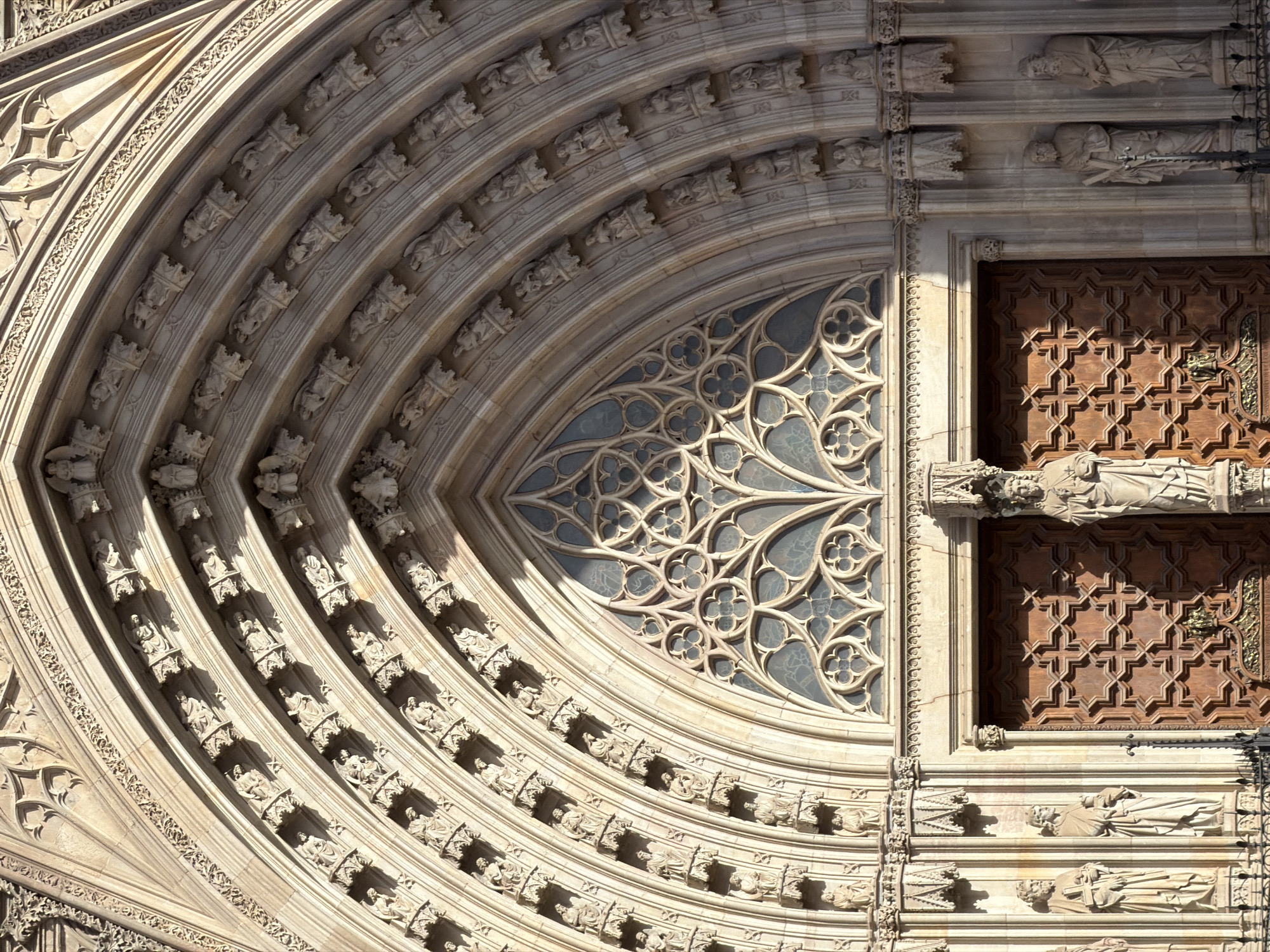 The image shows a close-up of the entrance to the Barcelona Cathedral, located in the Gothic Quarter of Barcelona, Spain. The entrance features a large, ornate wooden double door, intricately carved with a repeating geometric pattern. Above the door is a large, pointed arch with multiple recessed layers, each decorated with small sculpted figures. In the center of the arch, above the door, is a rose window with complex tracery, divided into smaller geometric shapes. Flanking the entrance are tall, narrow columns and niches, some containing life-sized stone statues of figures in flowing robes. The facade is made of weathered stone, with shadows cast by the sun highlighting the architectural details.