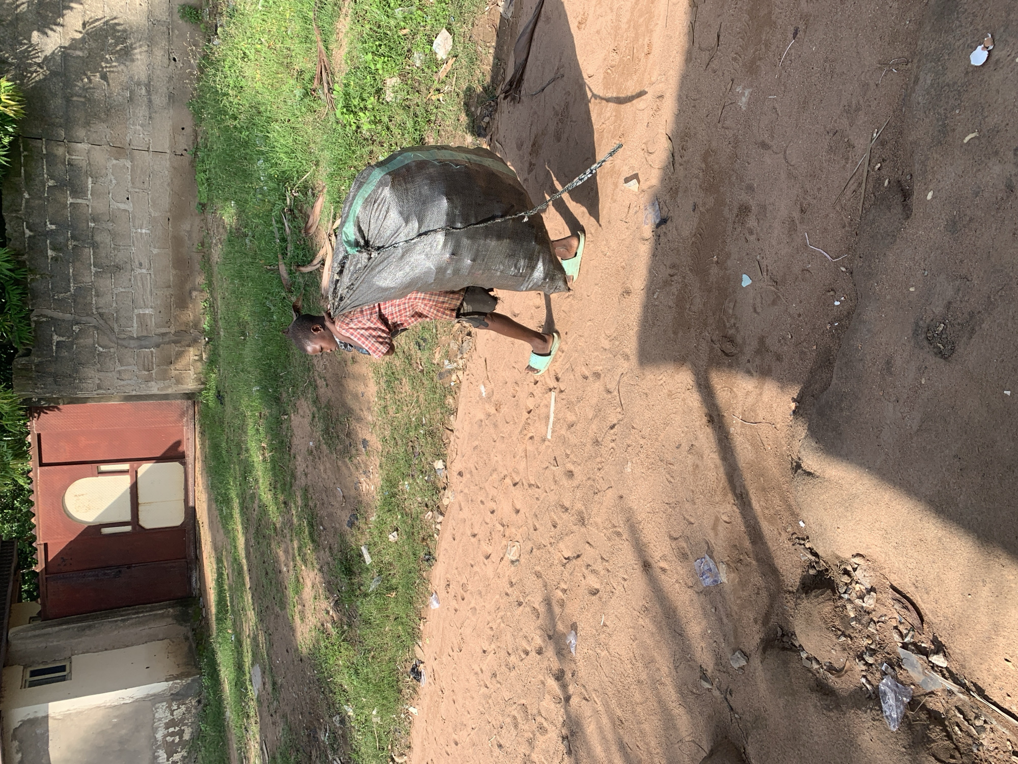 A young boy, appearing to be of African descent, walks across a dirt road carrying a very large, dark-colored sack of what appears to be trash over his left shoulder. He is wearing a red and white checkered shirt and dark shorts, with light blue flip-flops on his feet. The sack is almost as tall as he is and is secured with a rope or strap. The road is sandy and scattered with some debris and footprints. To the left of the road, there is a low-lying structure with a red gate and a small window, suggesting a residential or commercial building. Beyond that, there is a wall made of concrete blocks, overgrown with grass and some vegetation. The overall environment appears to be a rural or semi-urban area within Jalingo, Nigeria, characterized by its unpaved roads, simple structures, and abundant greenery. The lighting suggests it is daytime, with shadows cast by the boy and nearby foliage.