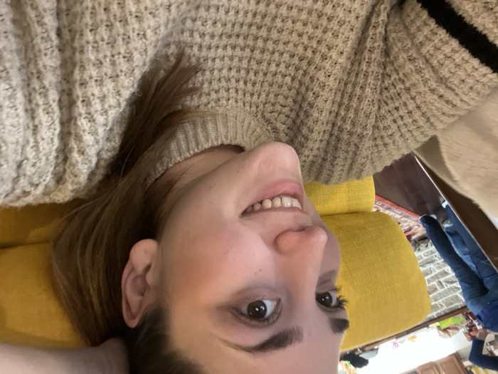 A female subject, appearing to be in her twenties, is depicted in a close-up, chest-up shot within an indoor residential setting in Ancenis, France. She has fair skin, light brown eyes, and light brown hair pulled back, with her right ear visible. Her head is angled towards the viewer's left, and she is smiling, revealing her upper and lower teeth. She is wearing a light beige or oatmeal-toned chunky knit sweater with two dark horizontal stripes visible on her left sleeve. Her left hand is partially visible, positioned behind her head. She is reclining against a bright yellow upholstered object, likely a couch or large cushion. In the blurred background to the left, the lower torso and legs of a second individual are visible, wearing blue jeans and a dark shirt, standing on a dark wooden floor with a red patterned rug. A light-colored wall with a stone or brick texture and a countertop with indistinct objects are also present in the background.