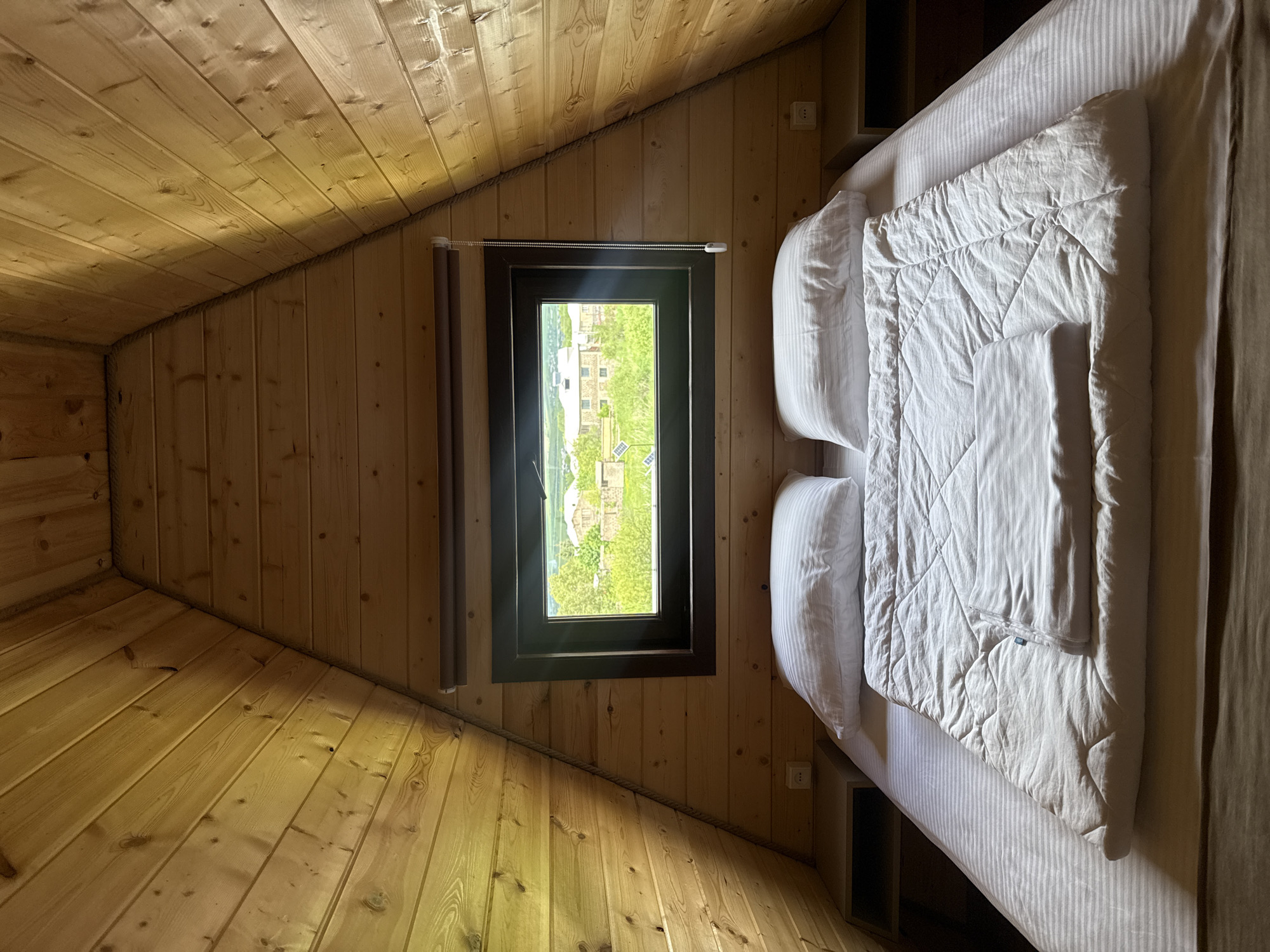 This is an interior shot of a room in a cabin in Stepanavan, Armenia. The room features wooden walls, ceilings, and floors, creating a rustic aesthetic. A bed with white linens, pillows, and a quilted blanket occupies the right side of the frame. A window with a black frame is centered in the image, providing a view of greenery and buildings outside. The perspective is slightly angled, emphasizing the sloping ceiling and triangular shape of the room. The room appears clean and well-maintained.
