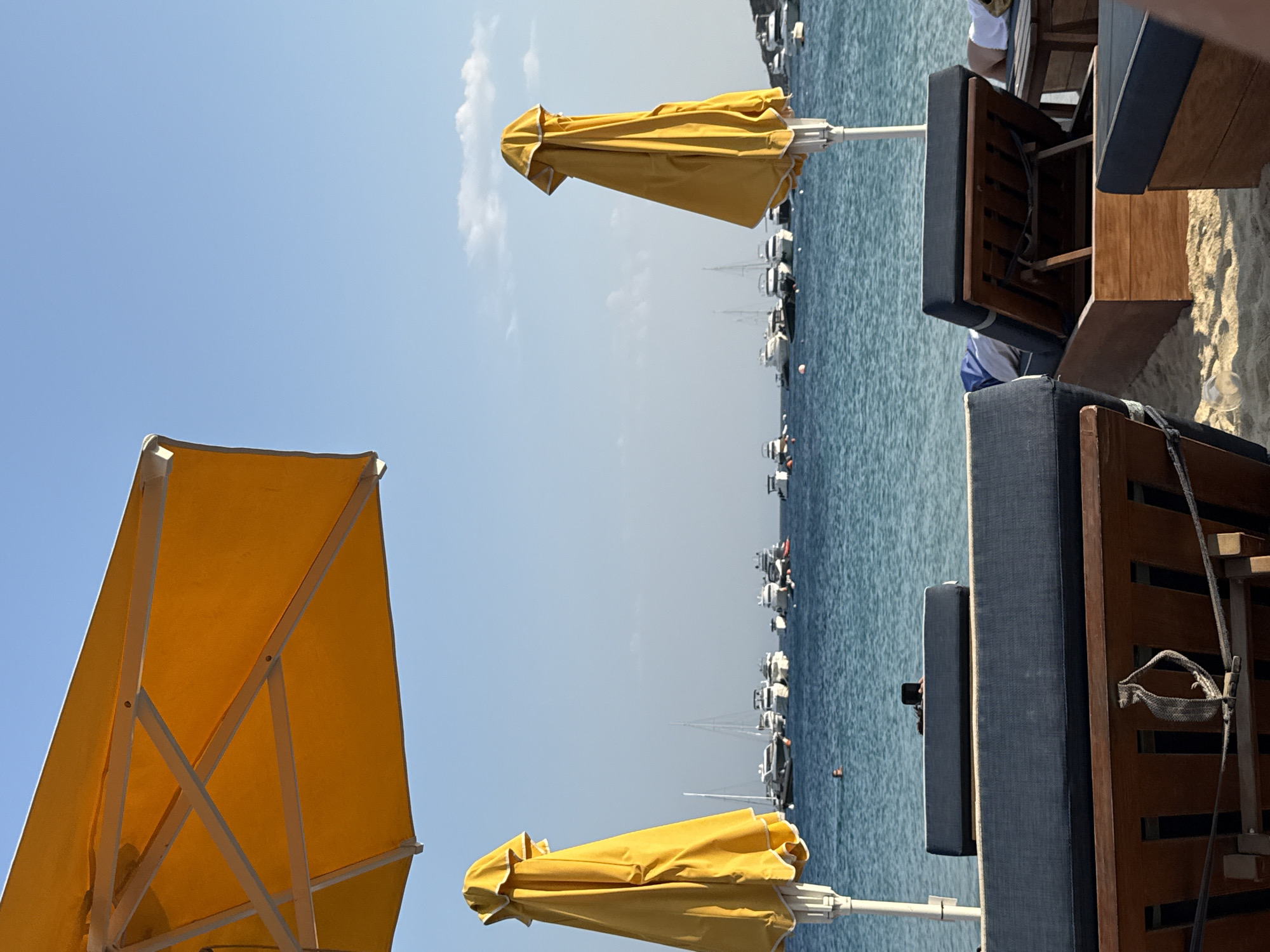 The image depicts a beach scene in Agios Ioannis Diakoftis, Greece, under a clear blue sky. In the foreground, two closed yellow beach umbrellas stand on poles, positioned to the left and right of the frame. The umbrella on the left is mostly visible from its underside, showing its white frame and yellow fabric. The umbrella on the right is fully visible, its yellow fabric gathered and secured. In front of the umbrellas, dark blue cushioned lounge chairs with wooden frames are arranged on the sandy beach. One chair is mostly visible in the lower left, and another is partially seen in the lower right, angled towards the sea.
The middle ground of the image features the turquoise waters of the sea. A few scattered white buoys are visible on the surface of the water. A person is seen swimming in the sea between the two umbrellas. In the background, a fleet of various white boats and yachts are anchored on the water, with their masts and superstructures clearly defined against the horizon. The sky above is a pale blue, with a few wispy white clouds scattered towards the right side of the image. The overall impression is a serene beach setting with amenities for relaxation and a view of docked vessels.
