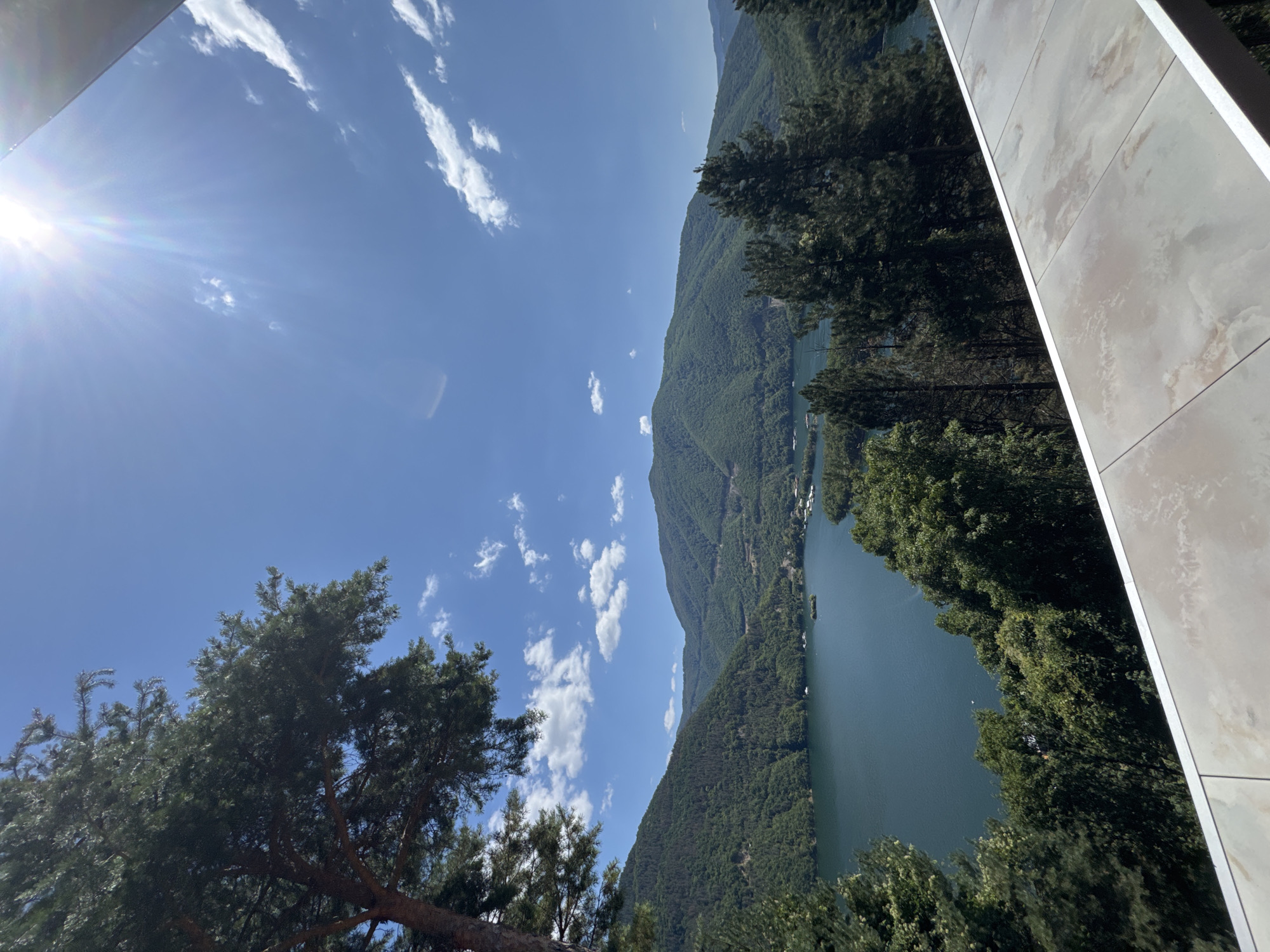 This is a high-angle, wide shot of a serene landscape featuring a body of water and surrounding mountains, captured on a bright, sunny day. The scene is likely situated in Mihalkovo, Bulgaria, a region known for its natural beauty.
In the foreground, the edge of a modern-looking balcony or patio is visible, with tiled flooring. A large, mature pine tree dominates the left side of the frame, its branches reaching towards the bright blue sky. The sky itself is clear with scattered white clouds and is illuminated by the intense sun, which creates a lens flare effect in the upper right portion of the image.
The middle ground is occupied by a vast, dark blue lake, nestled between rolling, densely forested green mountains. The mountains are covered in a thick canopy of trees, with no visible human structures apart from a few small buildings clustered on the far shore of the lake. Several small boats are scattered across the water, and a tiny island is visible in the lake. The overall impression is one of tranquility and natural splendor.