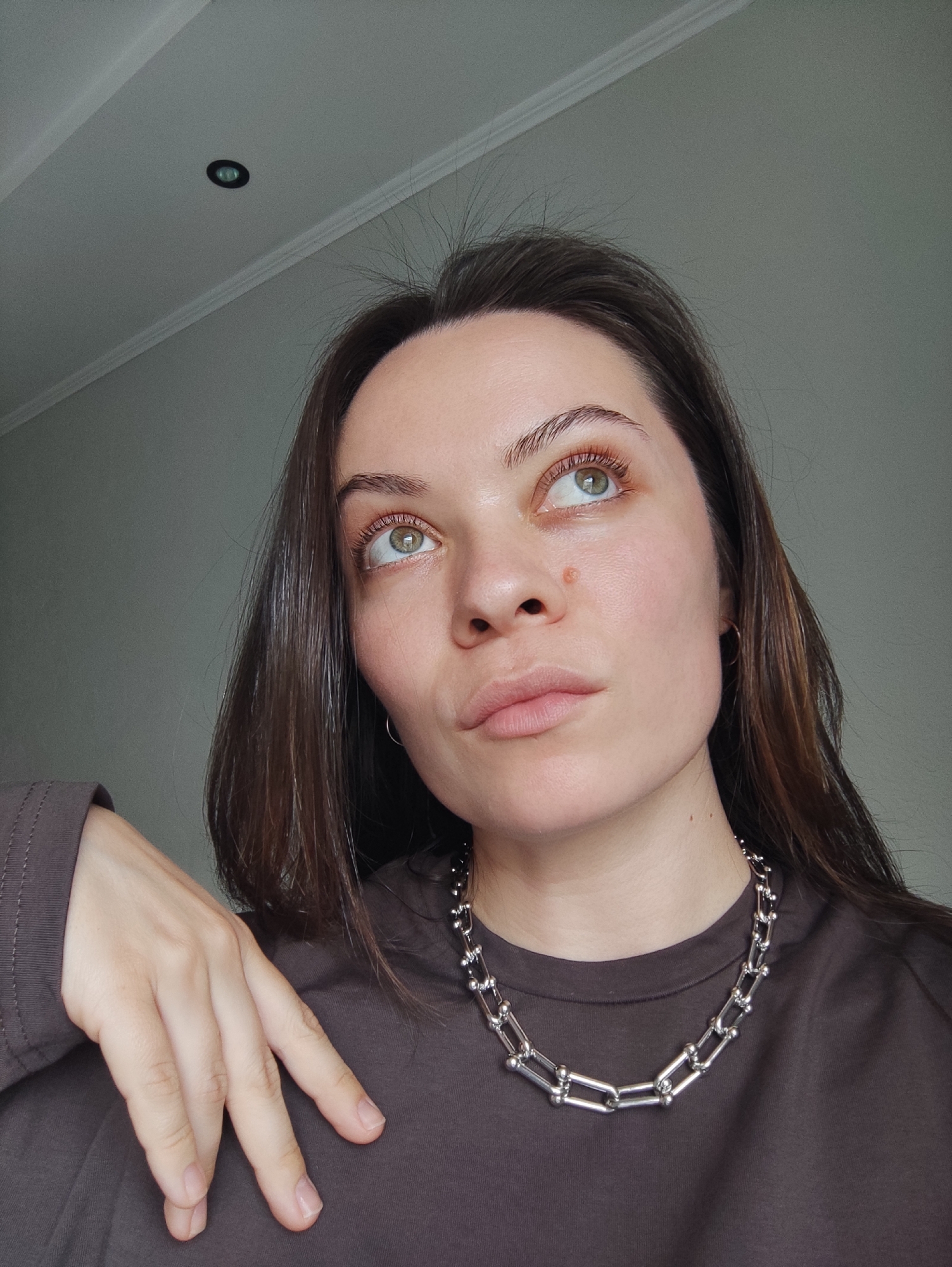 This media file is a portrait of a young woman, captured indoors from the chest up. She has long, dark brown hair, prominent green eyes, and fair skin, with a small mole visible on her nose. Her head is tilted slightly back, and she is gazing upwards and to her left with a thoughtful or contemplative expression.
She is wearing a plain, long-sleeved dark brown top and a distinctive chunky silver-colored chain necklace, composed of U-shaped links connected by spherical metallic beads. A small silver hoop earring is visible in her right ear. Her right hand, with neatly manicured nails, rests gently on her left shoulder or upper chest, partially obscuring her top.
The background consists of a plain light grey wall and a white ceiling featuring decorative molding and a single recessed circular spotlight. The lighting is soft and even, suggesting either natural daylight or diffused artificial illumination. There are no visible texts or specific details within the image to further identify the precise location beyond the general indoor setting in a modern space. The image is set in Chaikovsky, Russia.