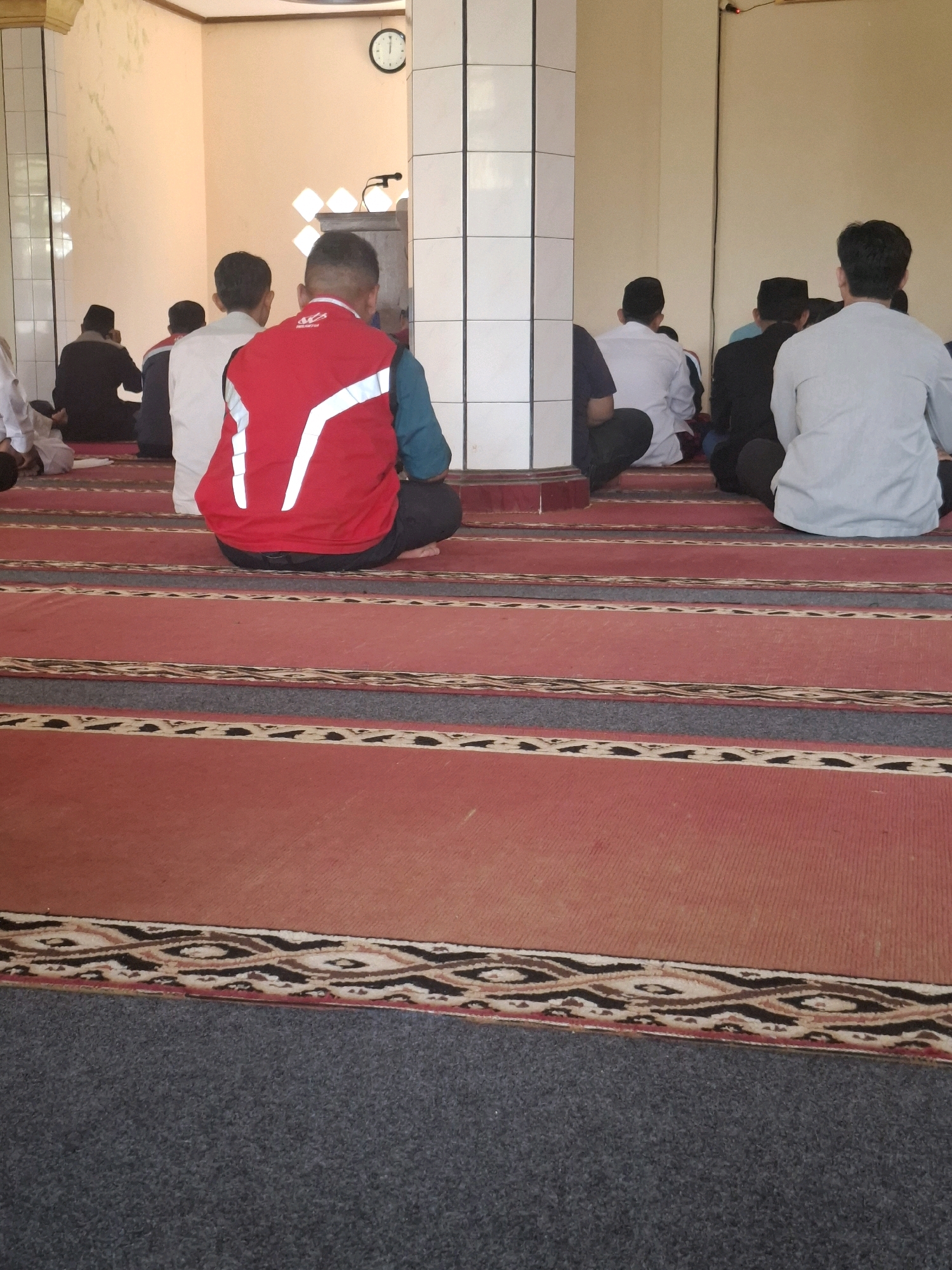 In a room with cream-colored walls and tiled pillars, several people are seated on a red carpet with patterned borders, facing away from the camera. A clock on the wall indicates it is past 12:00. Some individuals are wearing traditional Islamic attire, including white shirts and black skullcaps. One person in the foreground wears a red vest with white reflective stripes over a teal shirt and dark pants. The scene appears to be within a mosque or prayer hall, likely in Ciheulang Tonggoh, Indonesia, given the cultural context suggested by the attire and setting. The lighting is bright, suggesting daylight.
