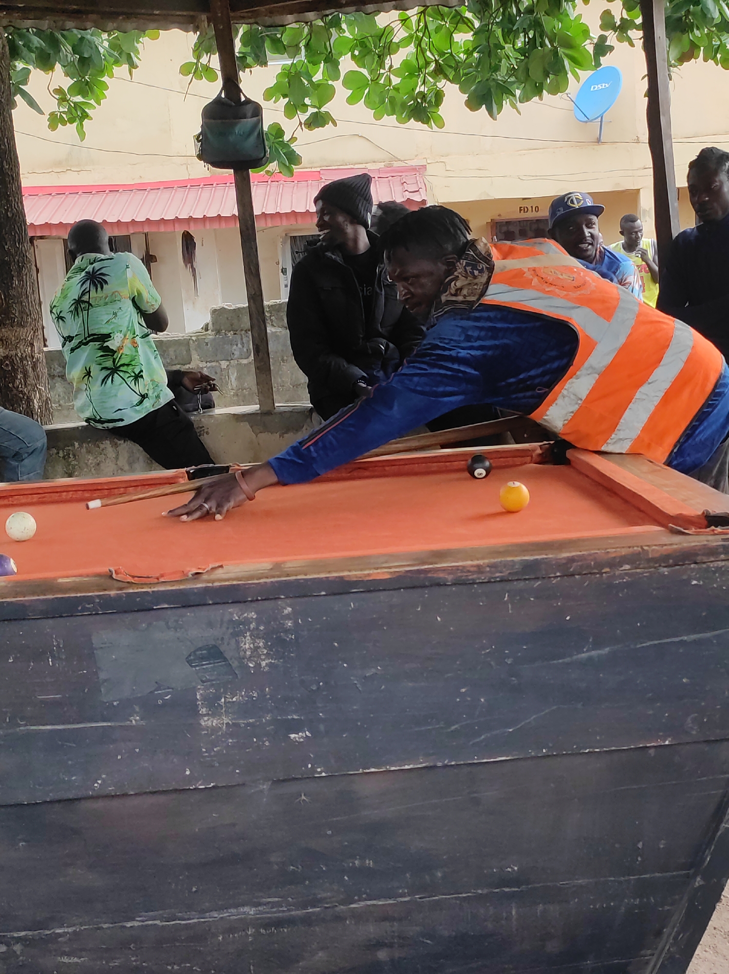 The image depicts an outdoor scene in Jos, Nigeria, where a group of men are gathered around a billiards table. A man wearing a blue jacket and a high-visibility orange vest is leaning over the table, cue in hand, preparing to strike a white cue ball. On the orange felt of the table, there is also a black eight-ball and a yellow ball. The billiard table is weathered, with a dark, worn base. Several other men are watching the game; one is wearing a light green shirt with a palm tree pattern, while others are wearing darker clothing and caps. In the background, there is a low concrete wall, foliage above, and a building with a red roof. A "DSTV" satellite dish is mounted on the building's wall. The scene appears to be casual and social.