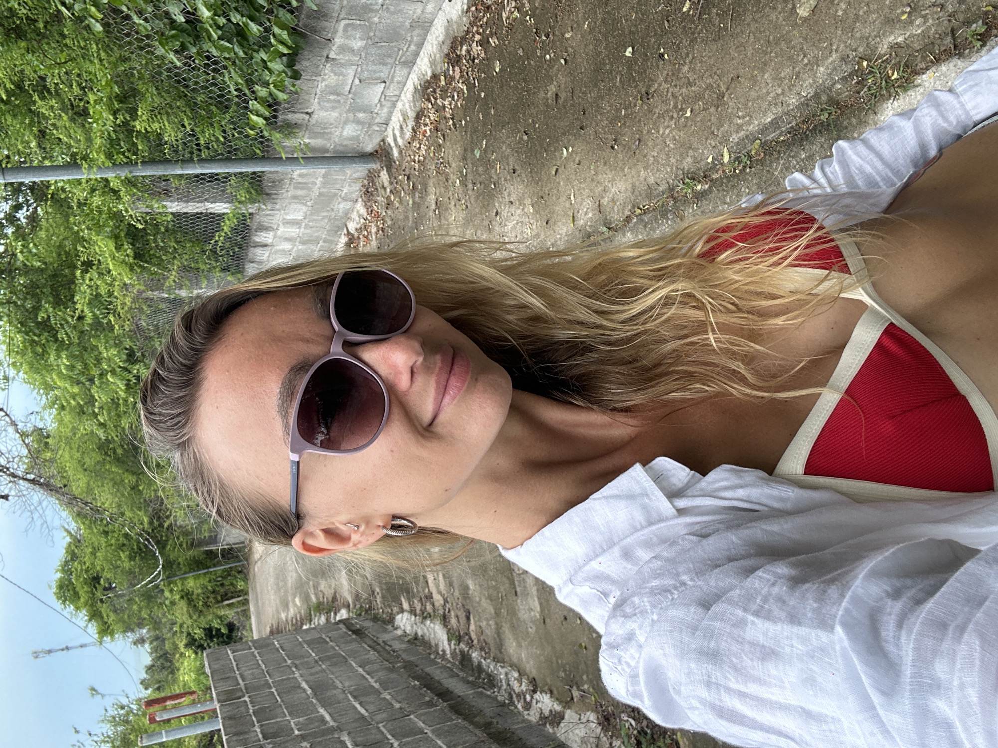 A woman with blonde hair and sunglasses smiles at the camera, taking a selfie outdoors. She is wearing a white button-down shirt over a red bikini top. The background shows a concrete pathway lined with a low brick wall and a chain-link fence, with lush green trees and vegetation beyond. The scene appears to be in Mũi Né, Vietnam, possibly near the coast or a residential area.