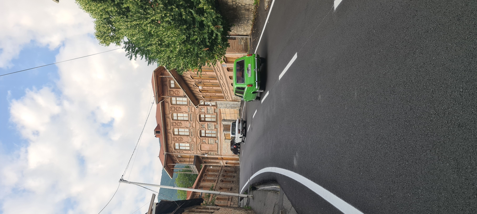 The image depicts a street scene in the city of Shaki, Azerbaijan, characterized by traditional architecture and a paved road. In the foreground, a newly paved asphalt road curves to the left, marked with crisp white lines. A bright green, older model car, possibly a Lada, is visible driving away from the viewer, with its license plate displaying "55 BA 221". Further up the road, a white car, also appearing to be an older model, is parked or driving slowly. Behind the white car, a dark colored car is also present.
The backdrop of the scene is dominated by ornate, multi-story buildings constructed with brick and featuring distinctive wooden balconies and intricate brickwork detailing. These buildings, typical of Shaki's historical architectural style, line both sides of the street, with one prominent building displaying "1893 F" inscribed above a window. Lush green trees flank the right side of the road, adding a touch of nature to the urban environment. The sky above is partly cloudy, with a mix of white clouds against a blue background. Utility poles and wires are also visible, indicating the presence of modern infrastructure. The overall impression is of a well-maintained street in a historically significant area.