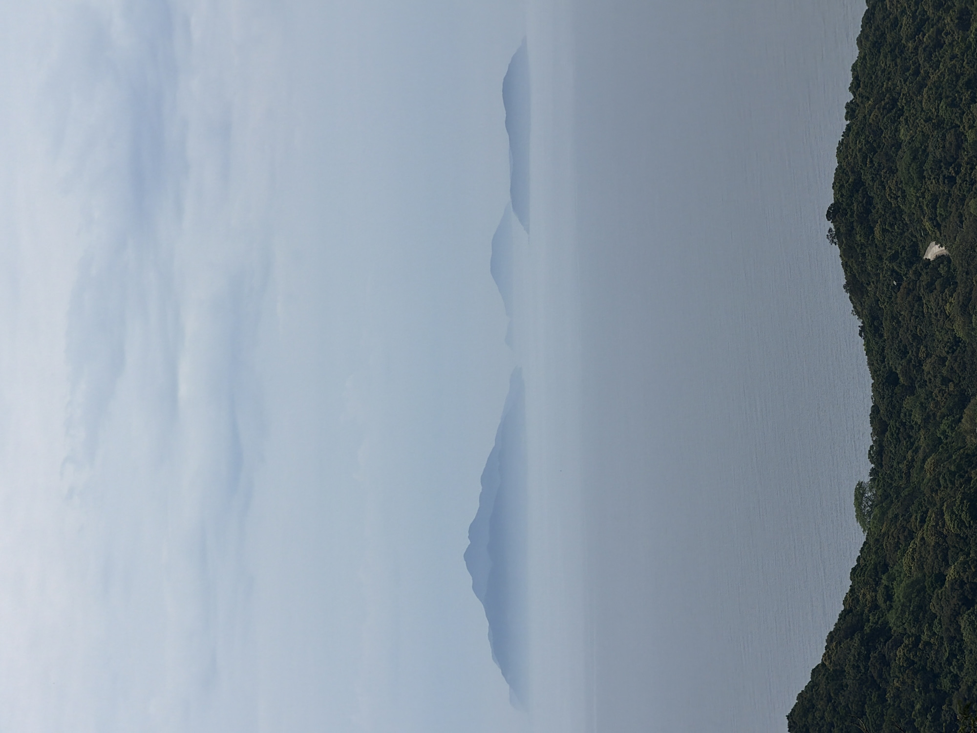 A view of the Sơn Trà Peninsula in Đà Nẵng, Vietnam, shrouded in mist. Lush, green vegetation covers the peninsula's steep slopes. Three peaks are faintly visible through the haze on the horizon, likely other islands or hills along the coast. The sea blends seamlessly into the misty sky.