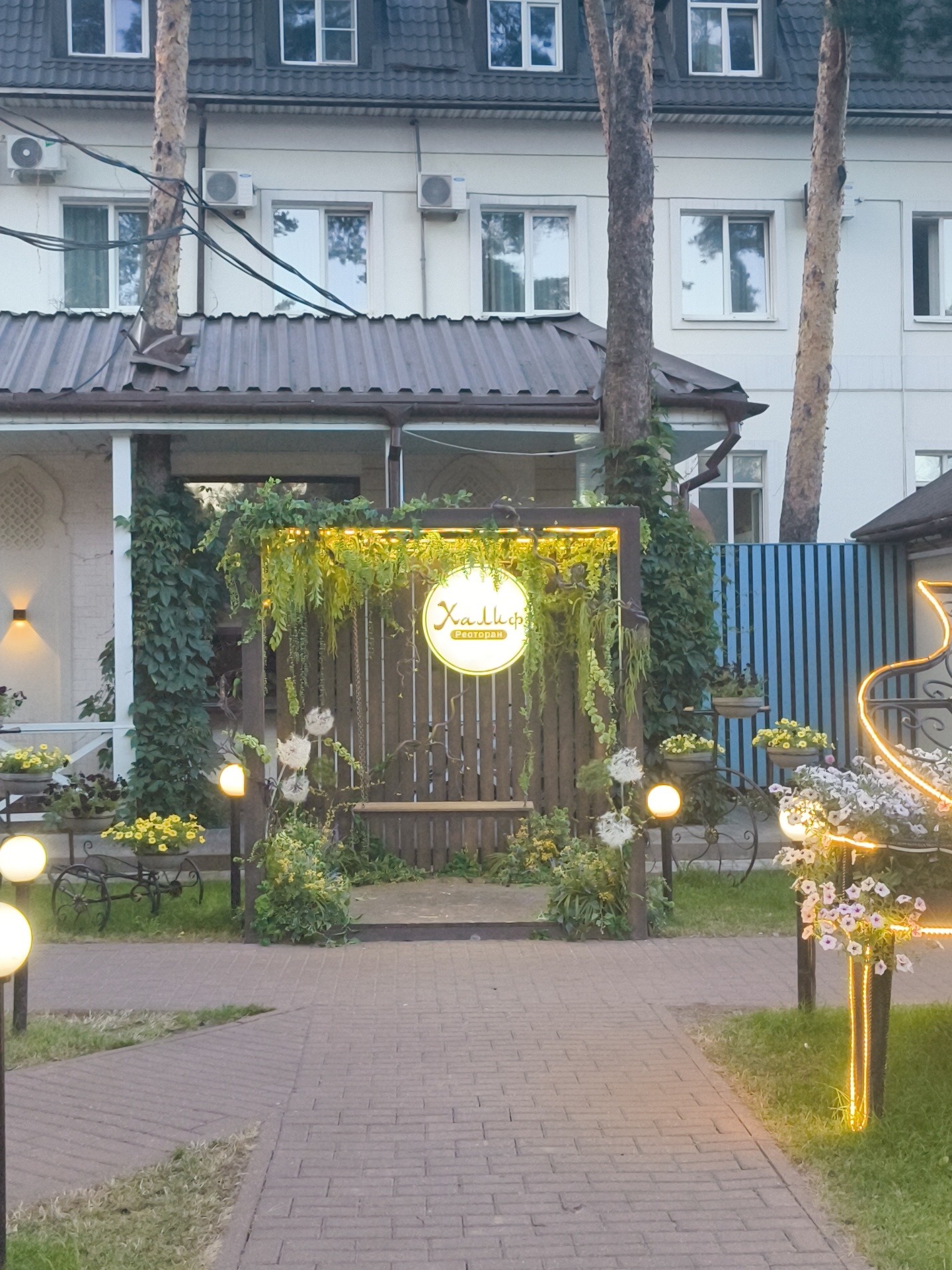 This is a medium shot, outdoor, eye-level photograph of an illuminated entranceway to a restaurant, likely in Ramenskoye, Russia. The entrance is decorated with greenery, hanging flowers, and a circular sign with Cyrillic text, possibly reading "Халифа Ресторан". The sign is lit from behind. A wooden bench is visible within the entranceway. The ground in front of the entrance is paved with brick. Several globe-shaped lights are placed along the pathway and within the decorated area. In the background, a building with multiple windows and air conditioning units is visible, with trees growing alongside it. To the right of the entrance, a blue corrugated metal fence is present. The lighting suggests it is dusk or evening, with artificial lights illuminating the scene. No people are visible.