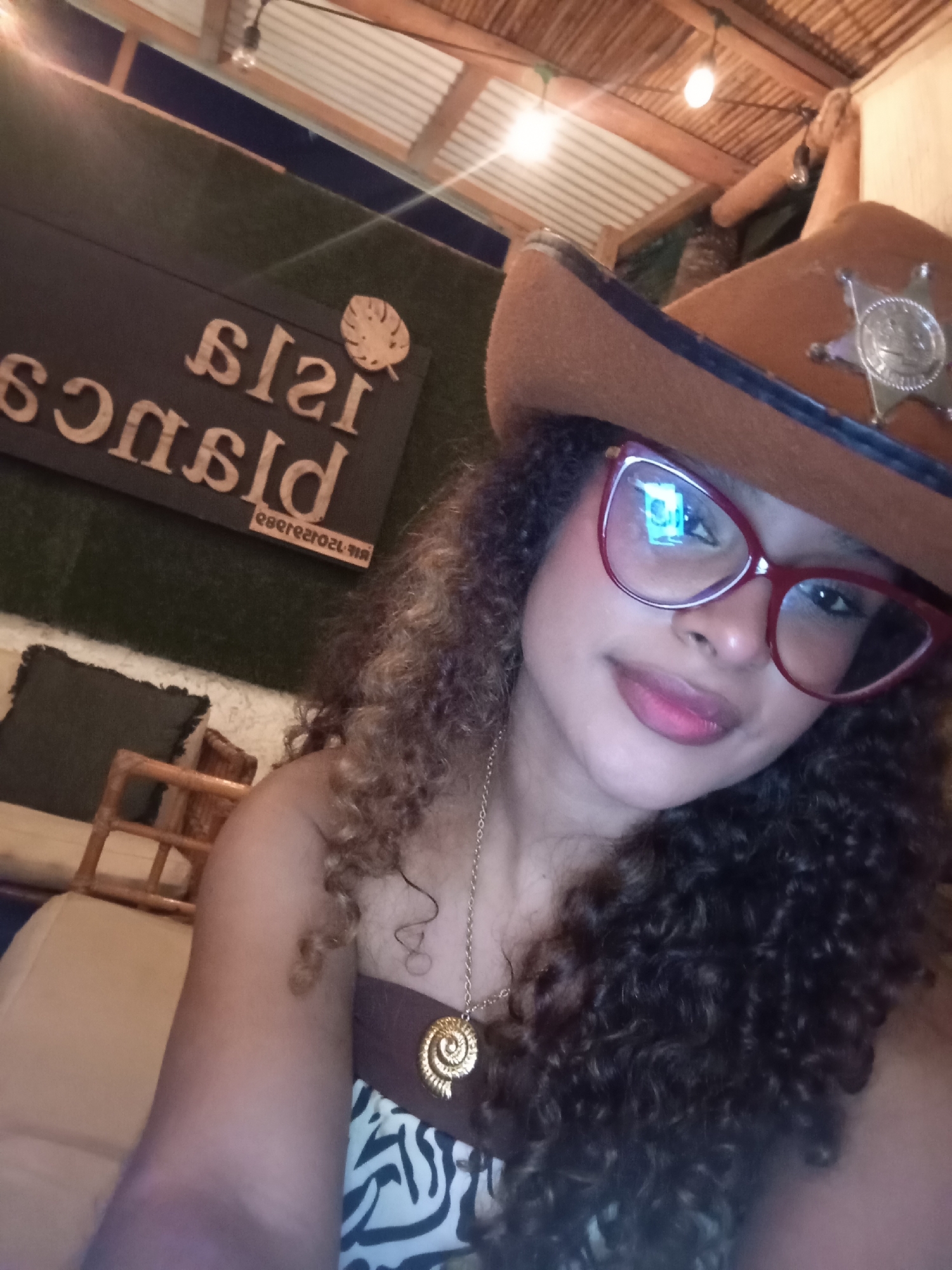This close-up selfie captures a young woman indoors, likely at a casual establishment in Porlamar, Venezuela. She has long, dark, curly hair and is wearing red-framed glasses that reflect ambient light. On her head, she wears a brown cowboy hat adorned with a silver sheriff's star badge. She sports a gold chain necklace with a unique spiraled shell pendant, and her outfit includes a strapless top with a brown band and a zebra print pattern. Her expression is calm, with a subtle, closed-lip smile.
The background reveals an indoor setting with warm lighting, suggesting it's evening or night. Above, a ceiling with corrugated panels, possibly bamboo or wood, and wooden beams is visible, from which string lights hang. To her left, a dark sign with light-colored, possibly wooden, reversed text is mounted on a wall covered in green artificial turf. When mirrored, the main text reads "ISLA BLANCA," with "RESTAURANTE" written in smaller letters below. Part of a seating area with a dark cushion and a light-colored wooden chair or bench frame is visible in the lower left.