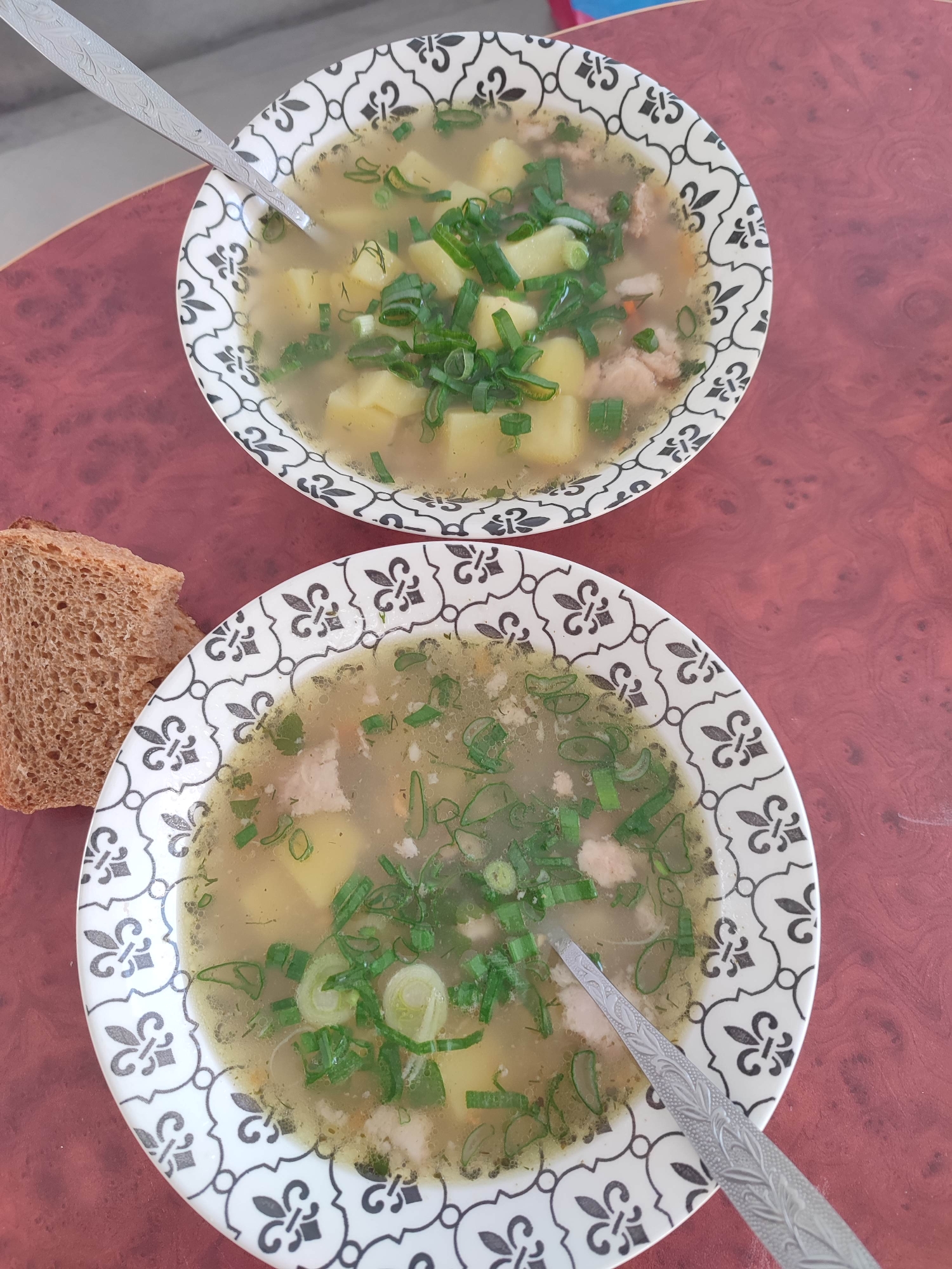 Two bowls of soup are placed on a reddish-brown table, with a slice of bread adjacent to the bottom bowl. Each bowl contains soup with visible ingredients, including potatoes, meat, and green onions, and a spoon rests inside. The bowls have a white base with a black fleur-de-lis-style pattern around the rim. The location is Unknown, Unknown.