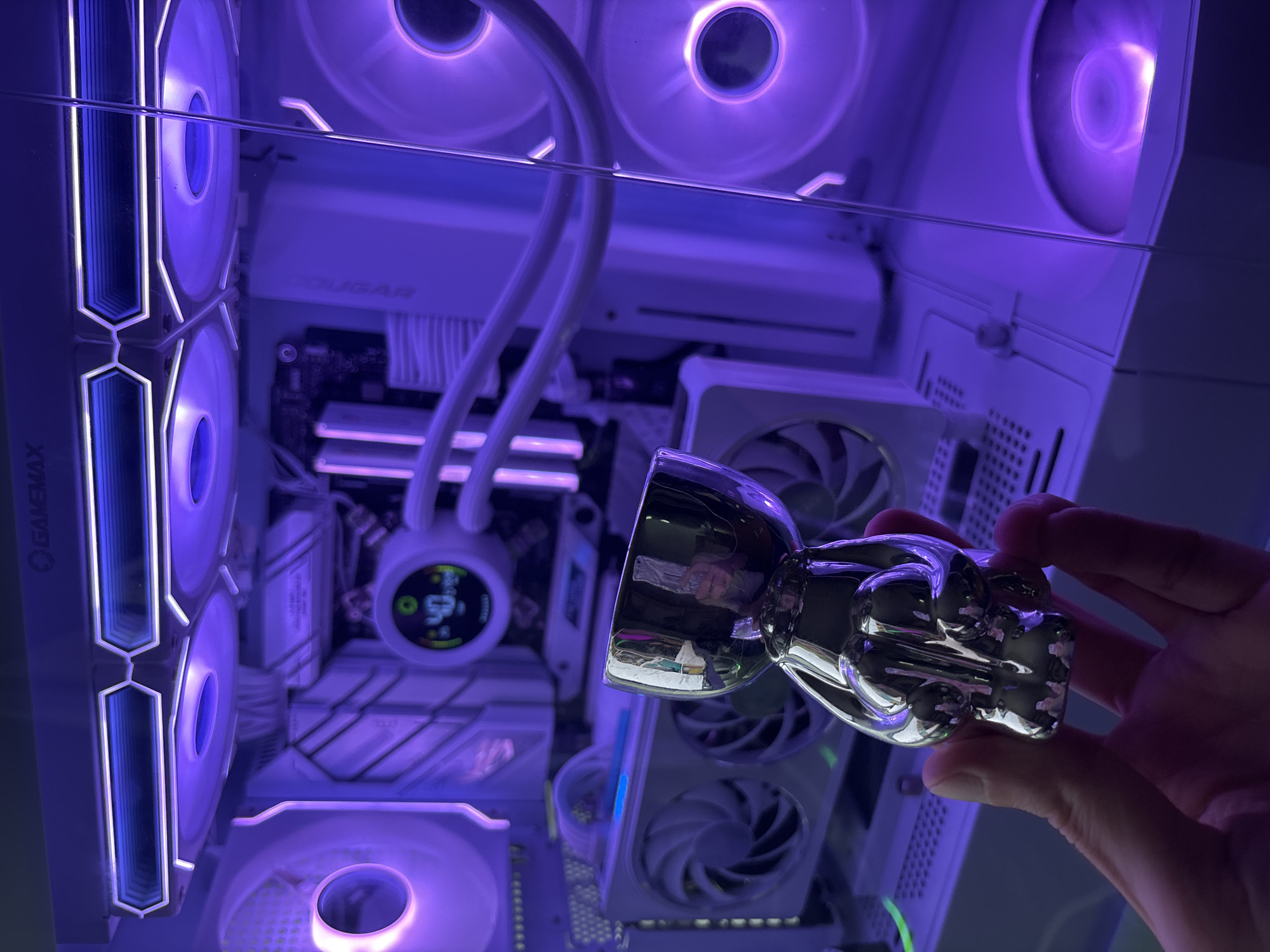 A person's hand is holding a small, shiny, silver skull-shaped object in front of a white computer tower with purple lighting. The computer tower has several fans and a liquid cooling system visible. The skull has a wide opening at the top, resembling a cup or chalice. The background is blurred, with the purple light illuminating the internal components of the computer.