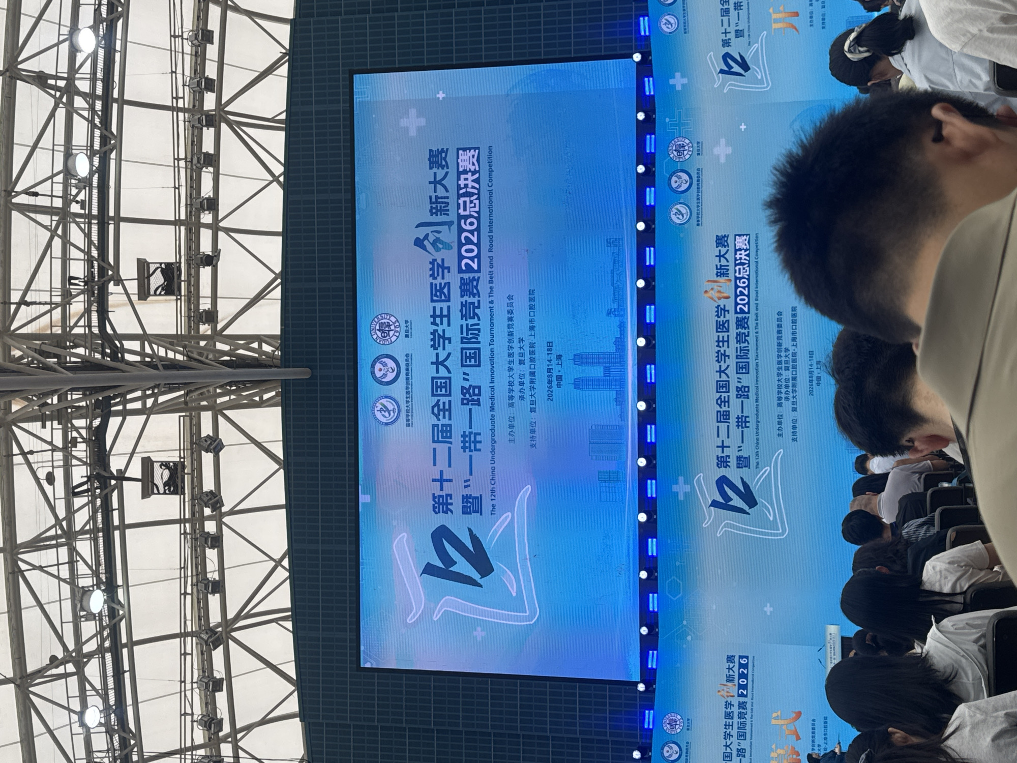 An overhead view captures a large screen displaying event details for "The 12th China Undergraduate Medical Innovation Tournament & The Belt and Road International Competition 2026." The screen, illuminated with a blue gradient and featuring prominent Chinese characters and logos, is set against a backdrop of stadium-like lighting and a structural ceiling. Below the screen, a crowd of people is seated, facing the display. The audience is largely obscured by the foreground, with only the backs of heads visible, suggesting a seated audience in a large venue. The overall impression is of a formal event or competition taking place in an indoor stadium or convention center. The location is identified as Yangpu District, China, indicating a specific geographical context for this event.