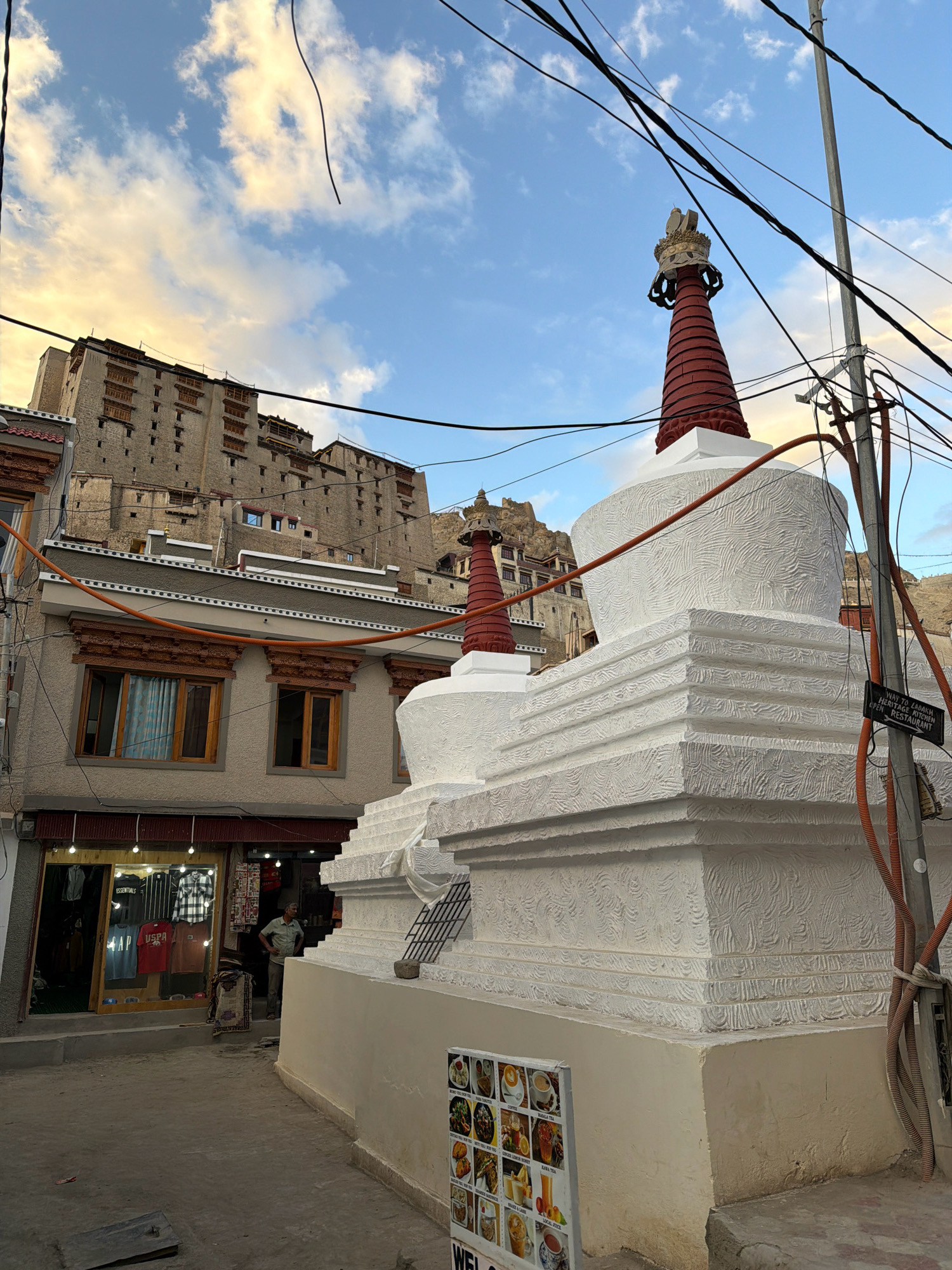 The image depicts a street scene in Leh, India, with traditional architecture and Buddhist stupas. In the foreground, two white stupas with red conical tops are prominently featured. The larger stupa on the right has intricate white patterns on its body and a tiered base. To the left of the stupas is a building with a storefront, displaying clothing items such as t-shirts and shirts. A man is standing near the entrance of the store, looking towards the right. Above this building, a multi-story structure with numerous windows, characteristic of Ladakhi architecture, rises up the hillside. The sky above is partly cloudy with hints of sunset colors. Electrical wires are visible crisscrossing the scene, suspended from poles. A sign attached to a utility pole on the right reads "WAY TO LADAKH HERITAGE KITCHEN OPEN RESTAURANT". The overall environment suggests a bustling urban area with a strong cultural identity.