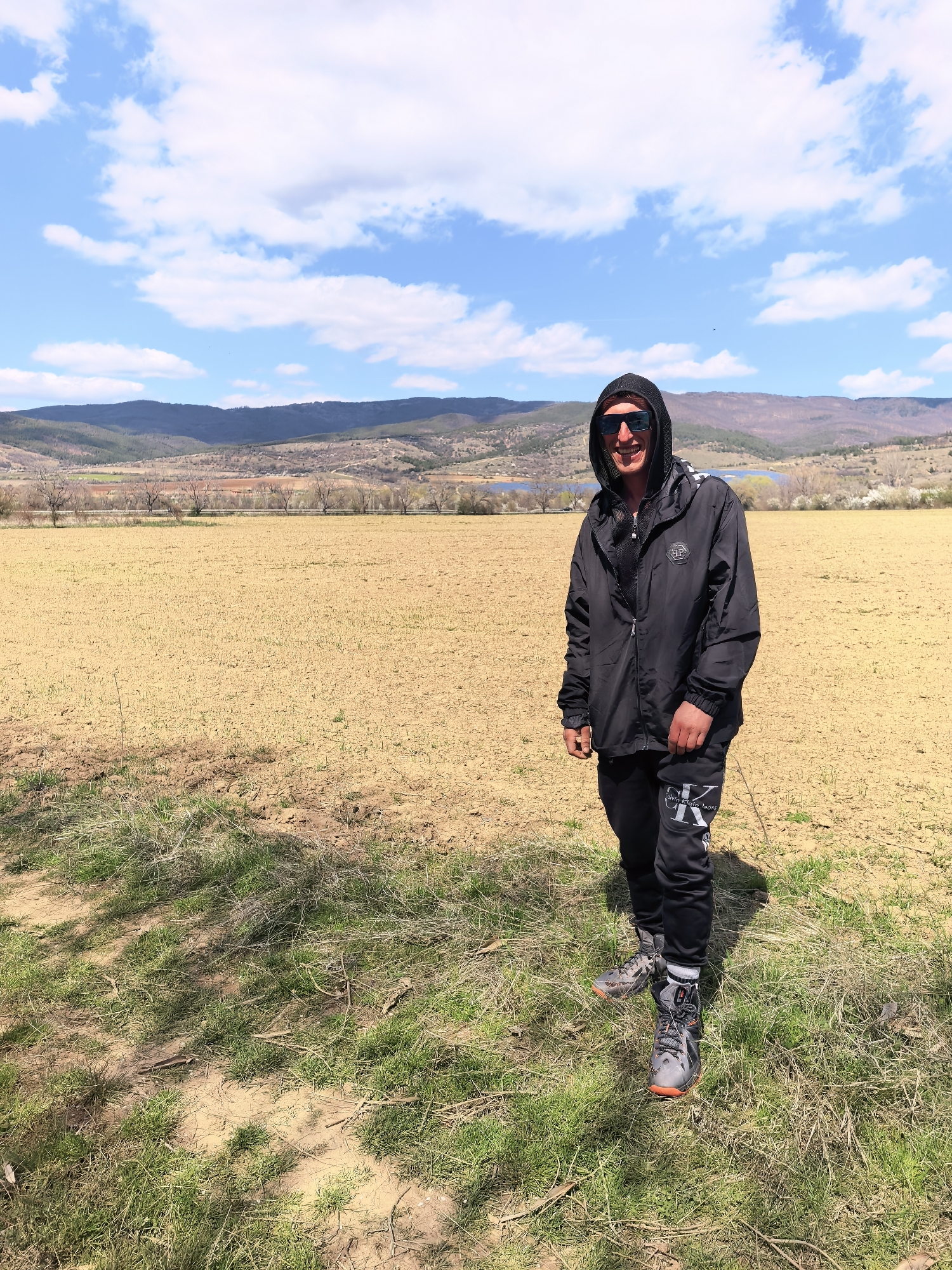 A man stands in a field of dry, light brown soil with some patches of green grass. He is wearing a black hooded jacket, black pants with a white "Calvin Klein Jeans" logo, and grey athletic shoes. Sunglasses are perched on his face, and the hood of his jacket is up. In the background, there are mountains, a blue lake, and a blue sky dotted with white clouds. The location appears to be a rural area.